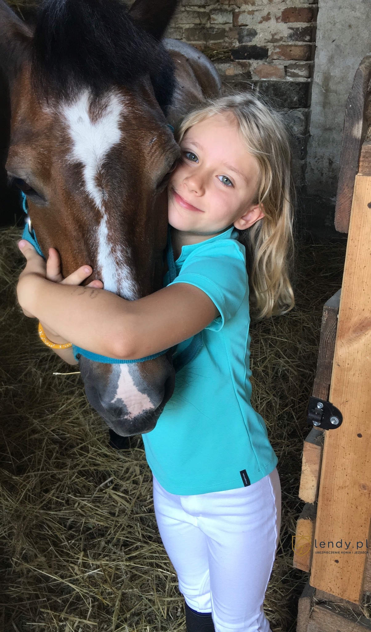 Dziewczynka w błękitnej koszulce i białych bryczesach przytula brązowego konia z białą łatą na czole w stajni.