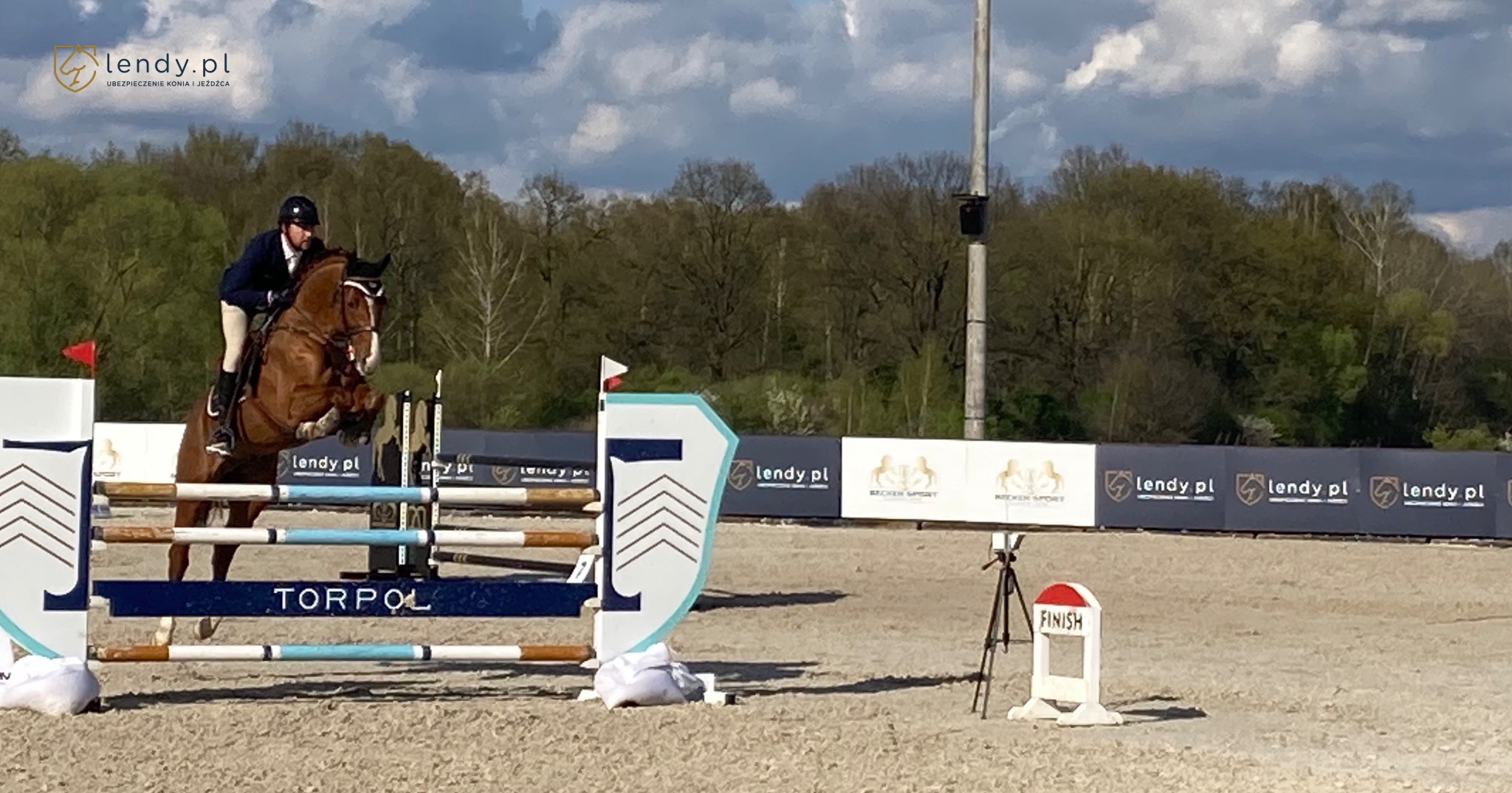 Jeździec i koń rasy kasztanowatej pokonują przeszkodę podczas zawodów jeździeckich na piaszczystym terenie, w tle widoczne drzewa i banery reklamowe z logo lendy.pl.