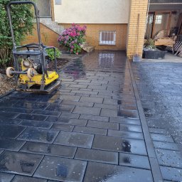 Adam Klimaszewski - Świeżo ułożona, błyszcząca, grafitowa kostka brukowa przed domem, z zagęszczarką w tle i fragmentem rabaty z hortensjami. Widoczny fragment garażu i wejścia do domu.