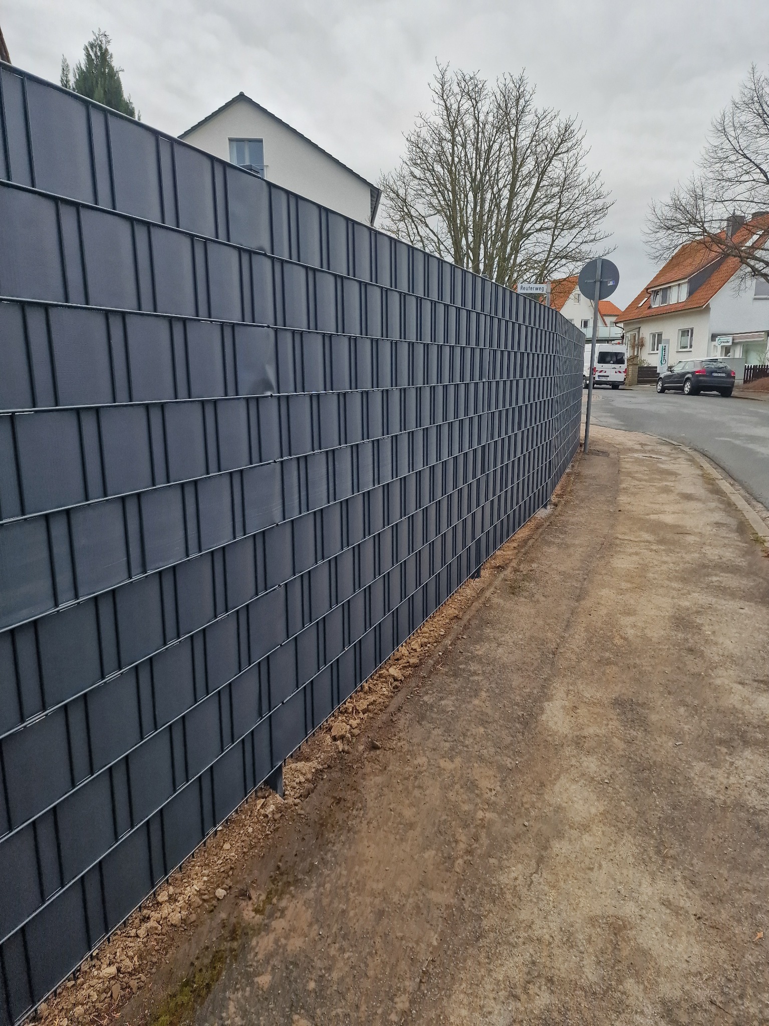 Nowoczesne, grafitowe ogrodzenie panelowe z widoczną strukturą, oddzielające posesję od chodnika i ulicy w zabudowie miejskiej. Widok z perspektywy przechodnia.