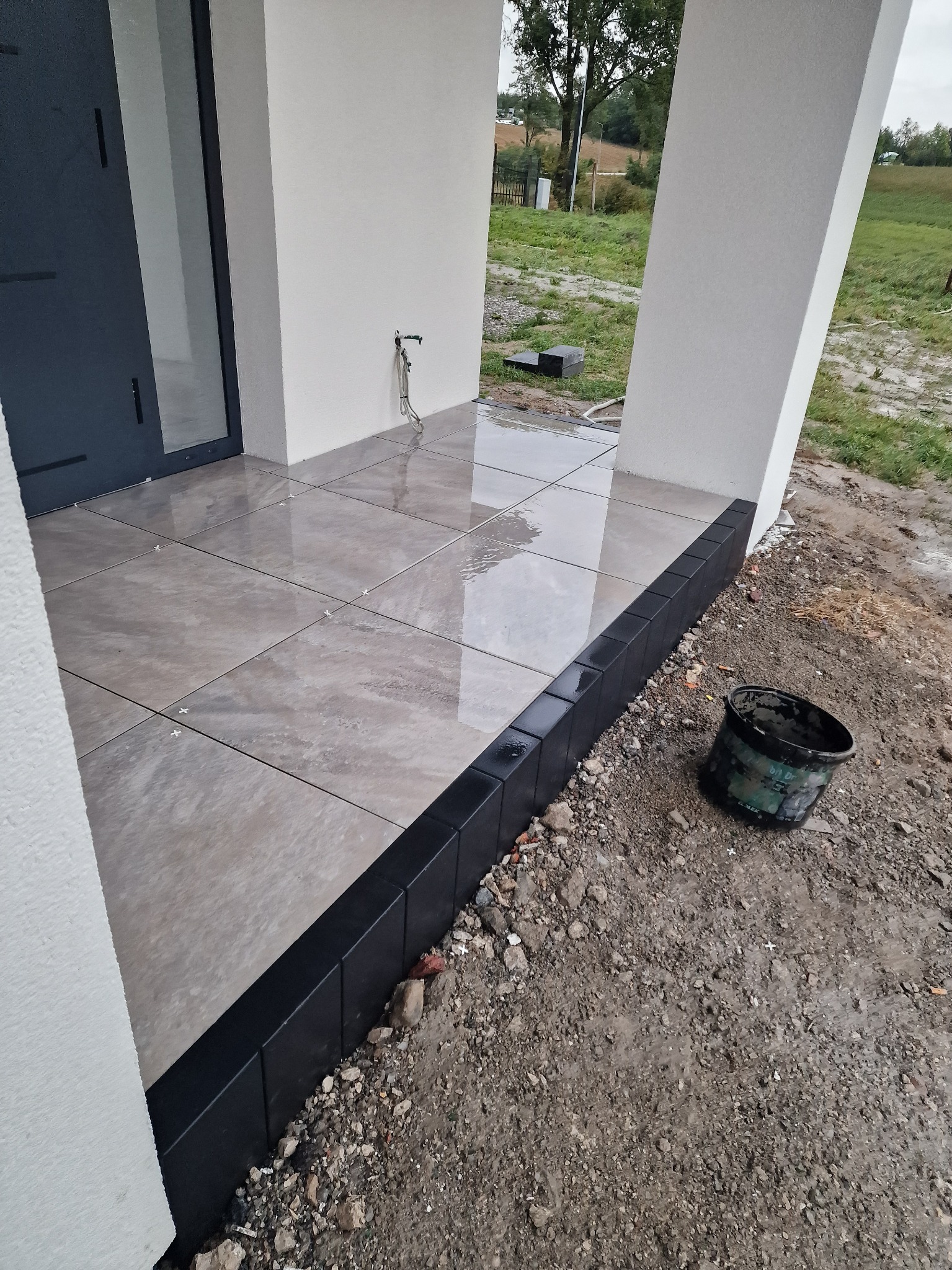 Wyłożony płytkami taras przy nowoczesnym domu z czarną obwódką. Widoczny kubeł budowlany i krzyżyki dystansowe. Ujęcie z niskiej perspektywy.