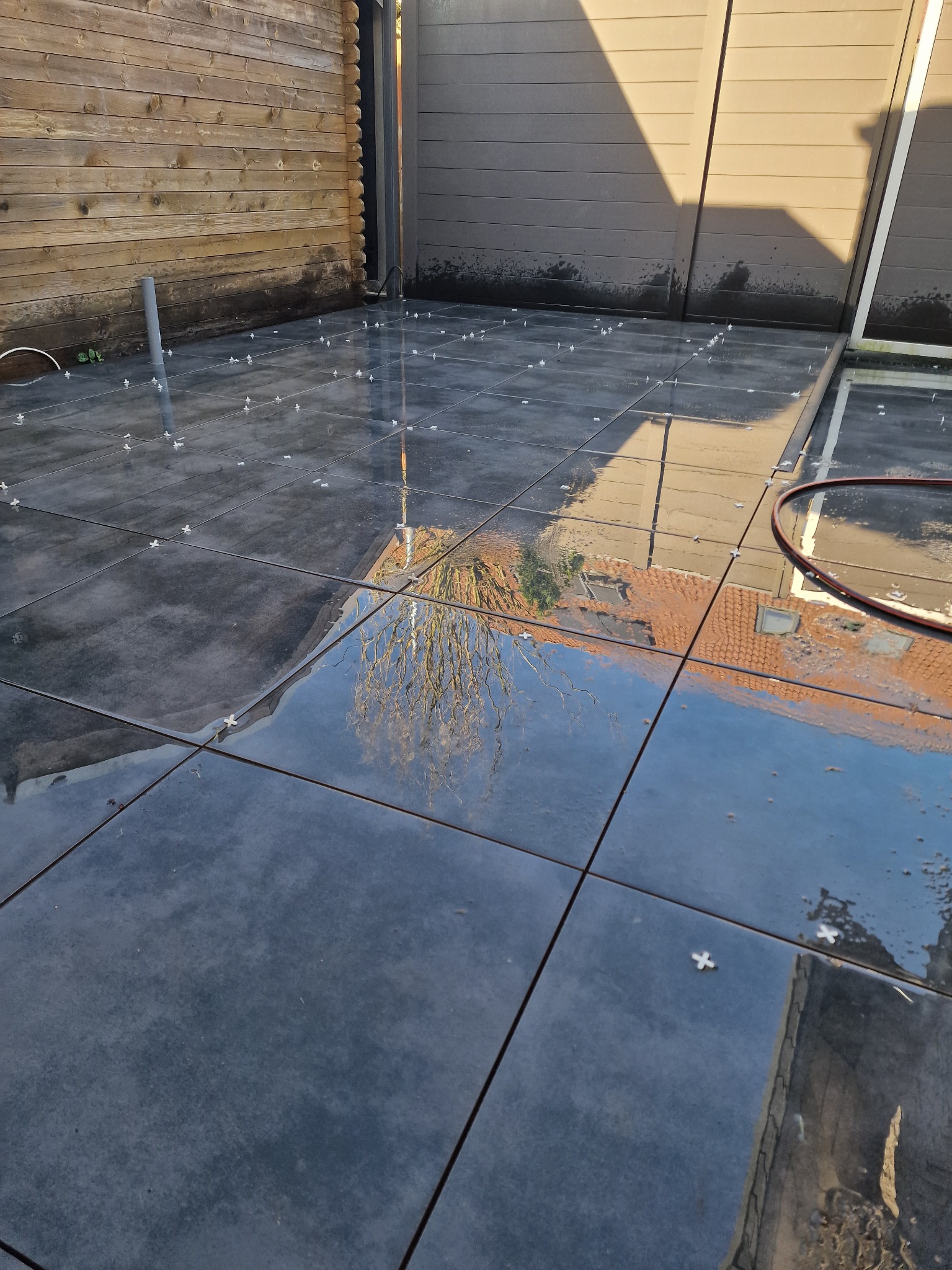 Błyszcząca, ciemna, kwadratowa płytka tarasowa w trakcie układania, z krzyżykami dystansowymi. Odbicie drzewa i budynku w mokrej powierzchni. Drewniana ściana w tle.