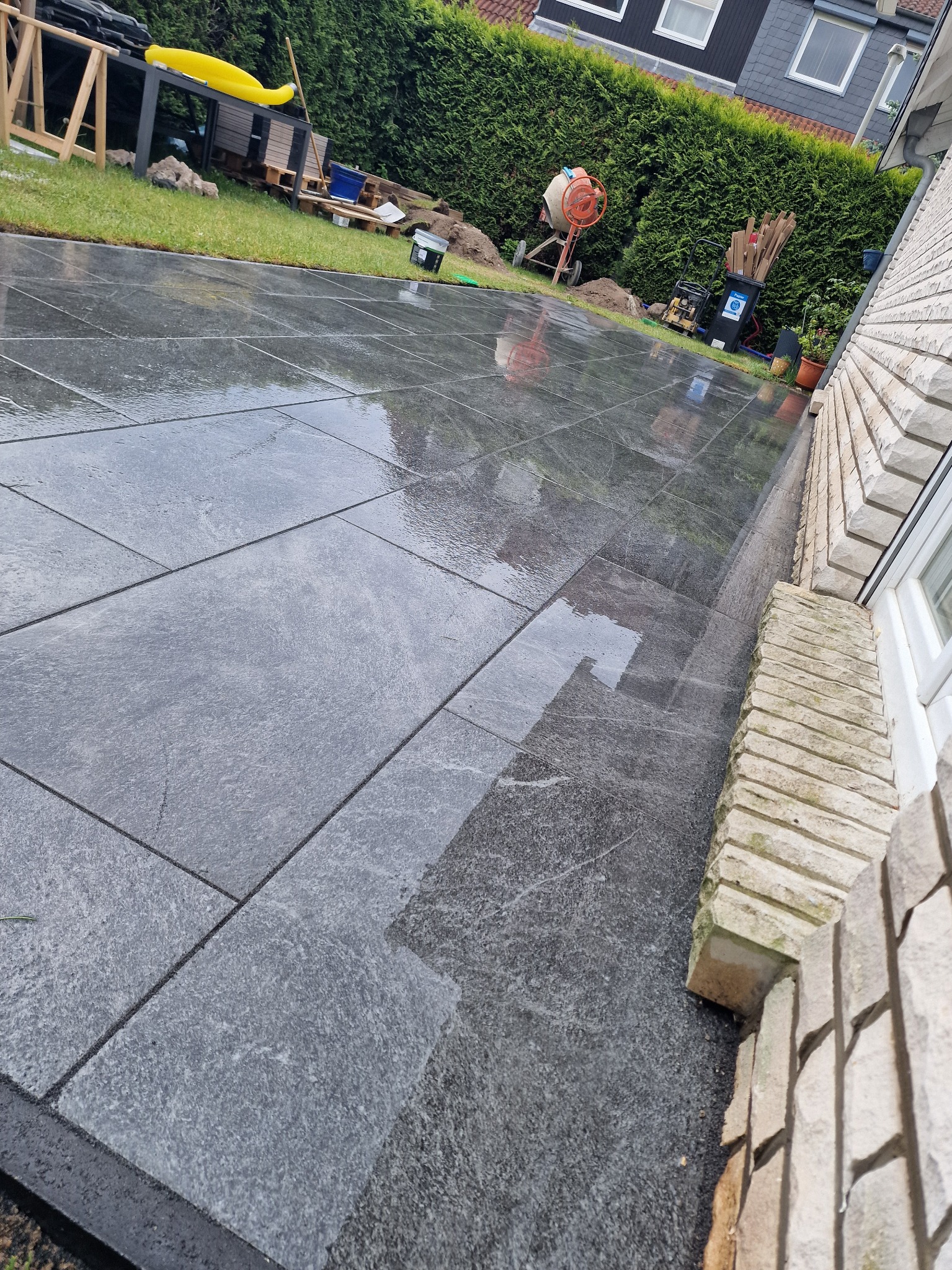 Świeżo ułożona, mokra, grafitowa kostka brukowa na posesji. W tle zielony żywopłot, sprzęt budowlany i fragment domu z jasnej cegły. Widok z góry, lekki kąt.