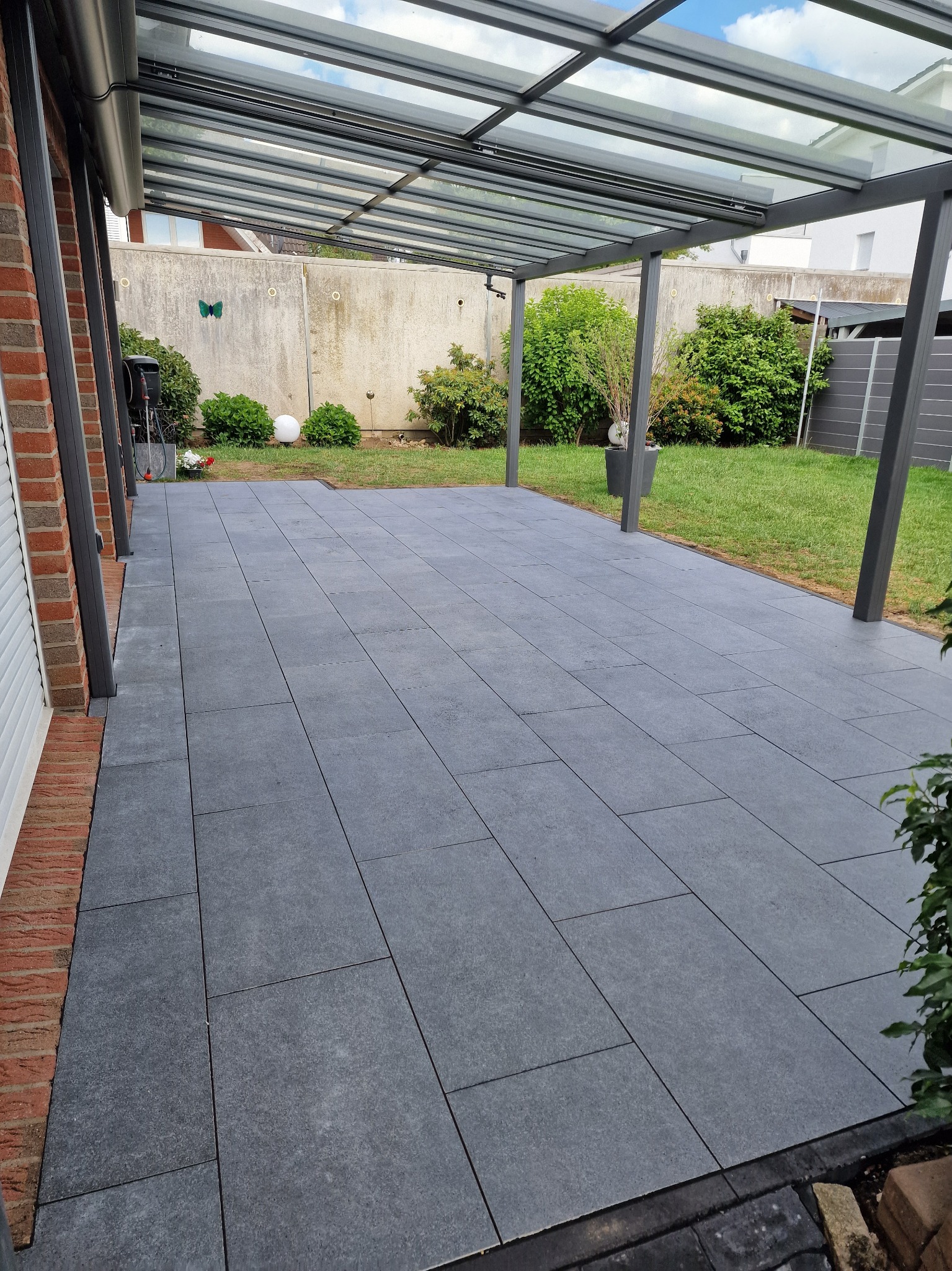 Nowoczesny taras z szarych płyt pod zadaszeniem, przylegający do budynku z ceglaną elewacją. Widoczne rośliny i trawnik w tle. Minimalistyczny design.