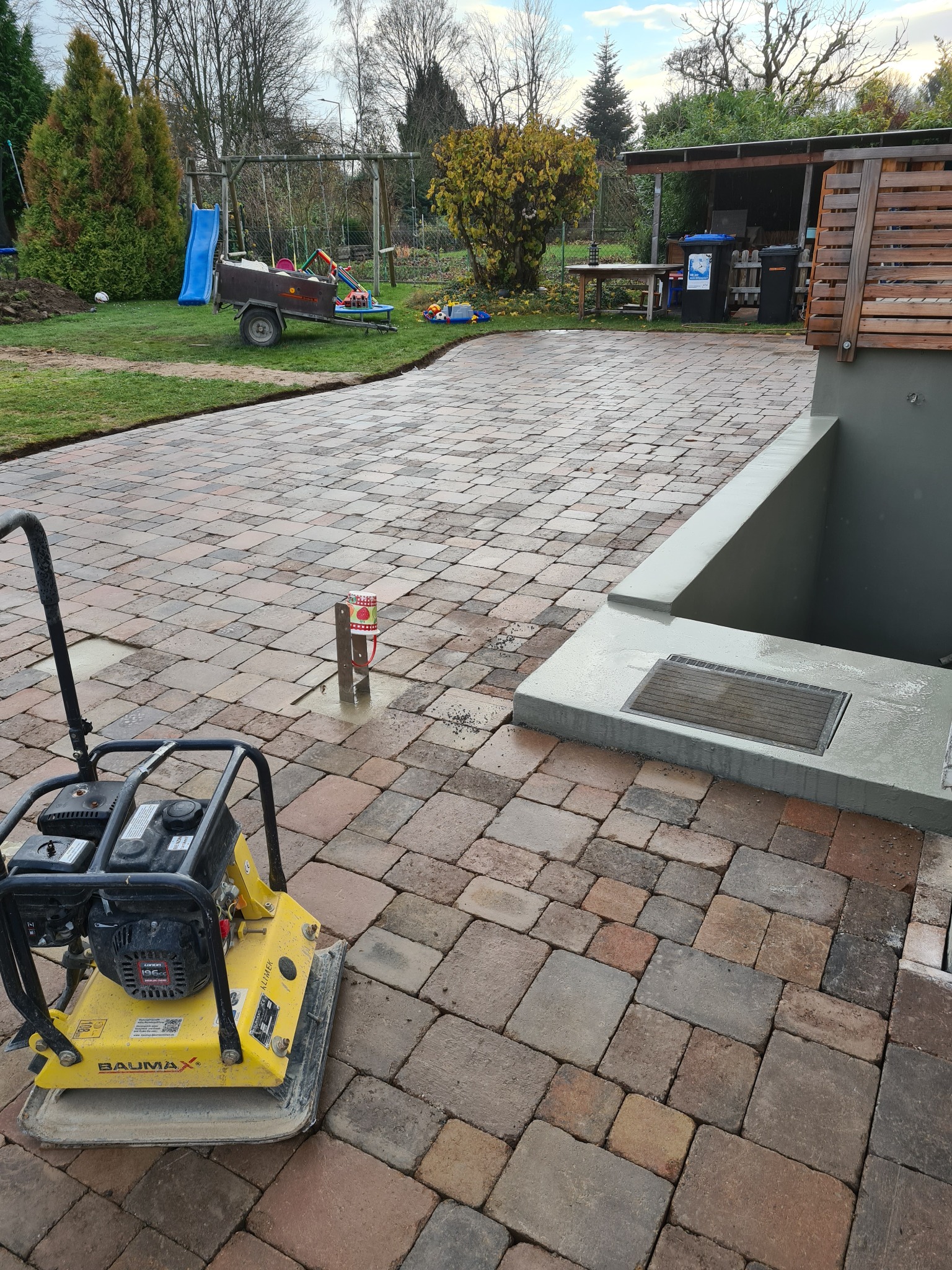 Świeżo ułożona, wilgotna kostka brukowa w odcieniach brązu i szarości, częściowo zagęszczona ubijarką wibracyjną BaumaX, z widocznym przyłączem wodnym i fragmentem budynku w tle.