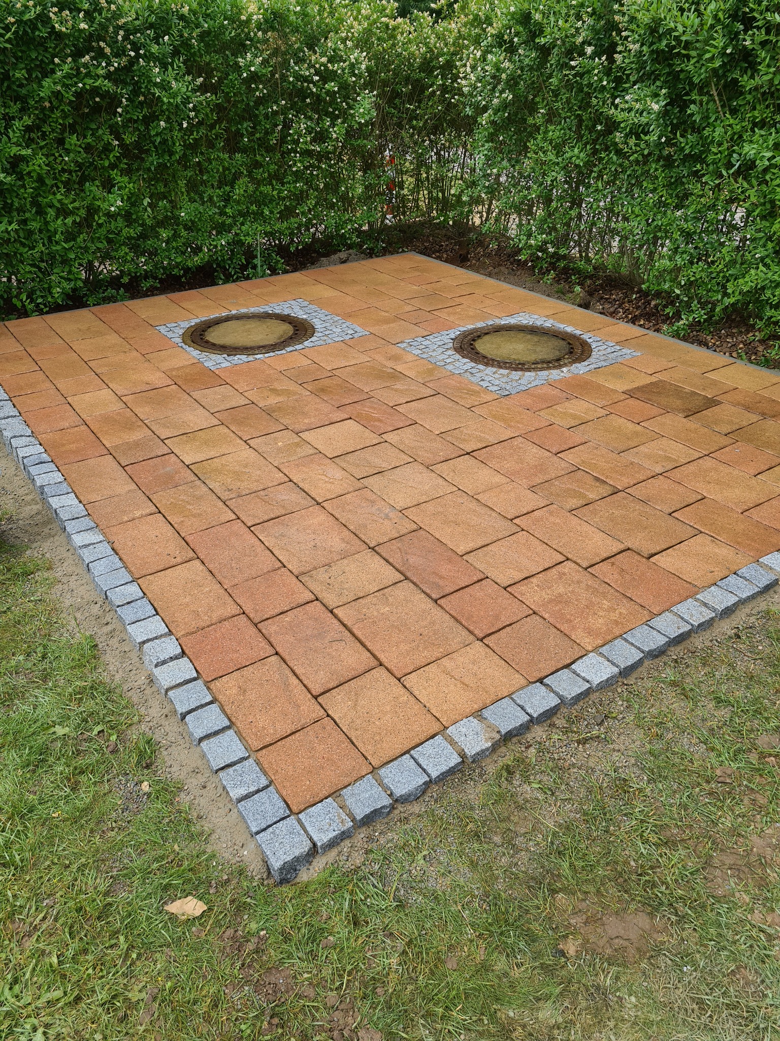 Ułożona kostka brukowa w kolorze pomarańczowym z szarym obramowaniem i dwoma włazami kanalizacyjnymi na tle zielonego żywopłotu.