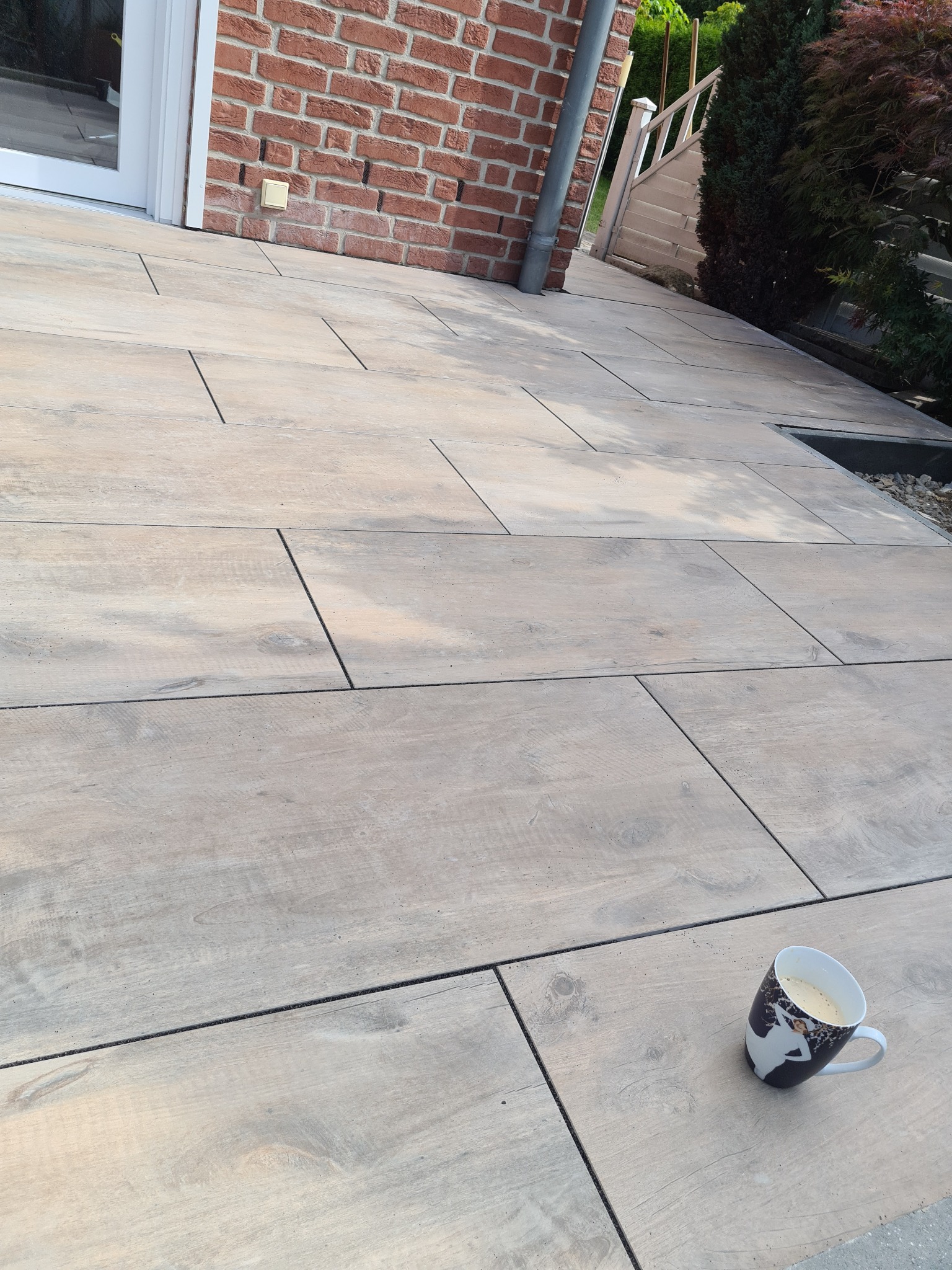 Nowoczesny taras z płyt imitujących drewno, z odpływem liniowym przy schodach i kubkiem kawy w rogu.