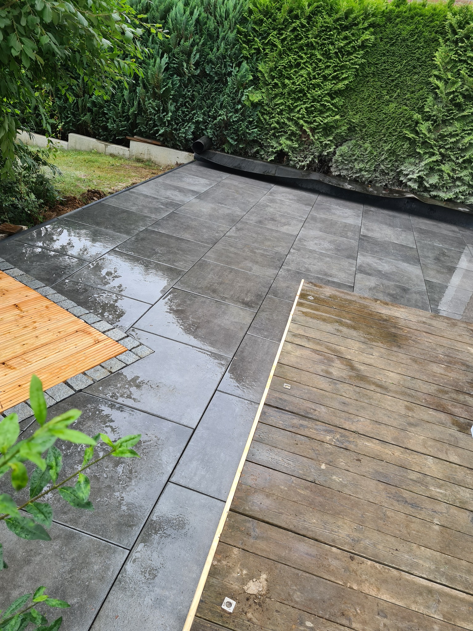 Świeżo ułożona, mokra, szara kostka brukowa w ogrodzie, sąsiadująca z drewnianym tarasem i obrzeżem z kostki granitowej, z zielonym żywopłotem w tle.