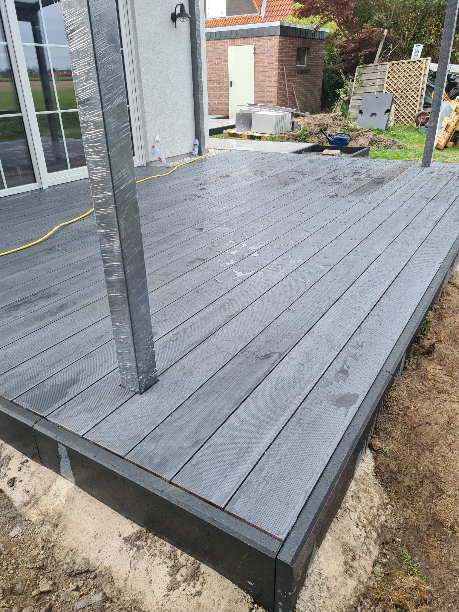 Nowoczesny taras z desek kompozytowych w odcieniach szarości, z widocznymi słupkami w folii ochronnej, w trakcie budowy, z ziemią wokół krawędzi i budynkiem w tle.
