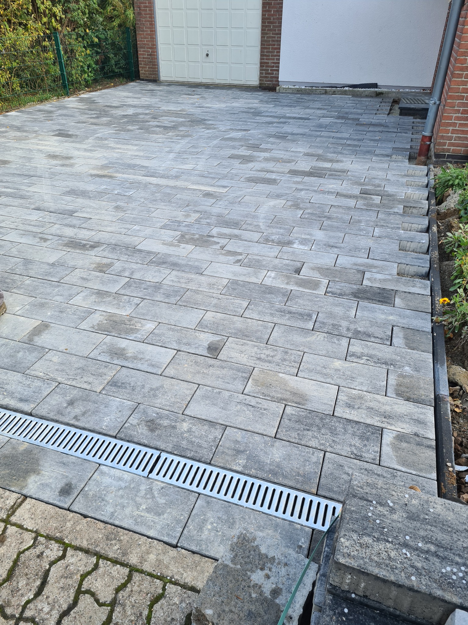 Ułożona szara kostka brukowa na podjeździe, z widocznym odpływem liniowym ze stali nierdzewnej, prowadząca do garażu z bramą segmentową.