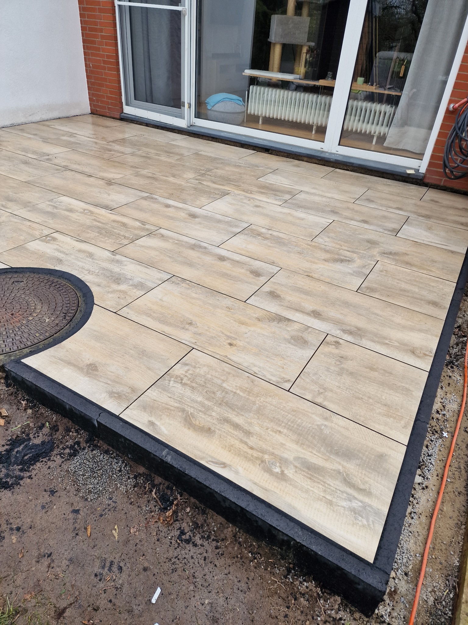 Nowy taras wyłożony jasnymi płytami imitującymi drewno, z czarną obwódką i studzienką kanalizacyjną w rogu, widok z góry na przesuwną szklaną ścianę domu.