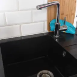 Czarny, granitowy zlew kuchenny z chromowaną baterią, drewnianą deską do krojenia i turkusową ściereczką w tle, pod białymi kafelkami.