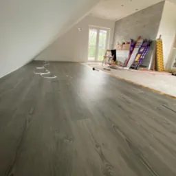 Szare panele podłogowe ułożone w nowym pokoju na poddaszu, widoczne narzędzia i materiały budowlane w tle.