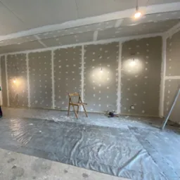 Montaż płyt kartonowo-gipsowych na ścianach i suficie, widoczne białe łączenia, podłoga zabezpieczona folią, składane krzesło na środku pomieszczenia.