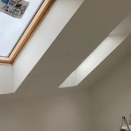 Świeżo wyszpachlowane poddasze ze skośnym sufitem, widoczne dwa okna dachowe Velux, jedno z naklejoną gazetą reklamową, aluminiowa drabina oparta o ścianę, ślady po gniazdkach elektrycznych.