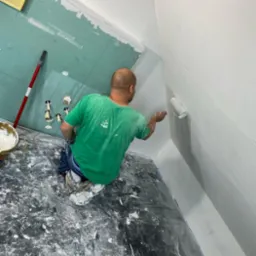 Mężczyzna w zielonej koszulce maluje białą farbą ścianę z płyt kartonowo-gipsowych za pomocą wałka, widoczne ślady farby na podłodze zabezpieczonej folią.