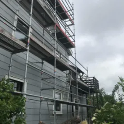 Dom jednorodzinny z szarą elewacją, otoczony rusztowaniem, przygotowywany do prac elewacyjnych w pochmurny dzień.