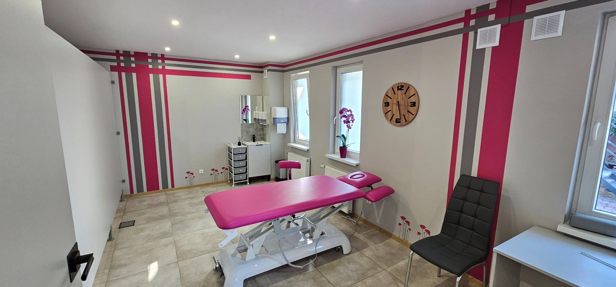 Jasne wnętrze gabinetu z różowym stołem do zabiegów, umywalką, drewnianym zegarem i dekoracyjnymi kwiatami na ścianach.