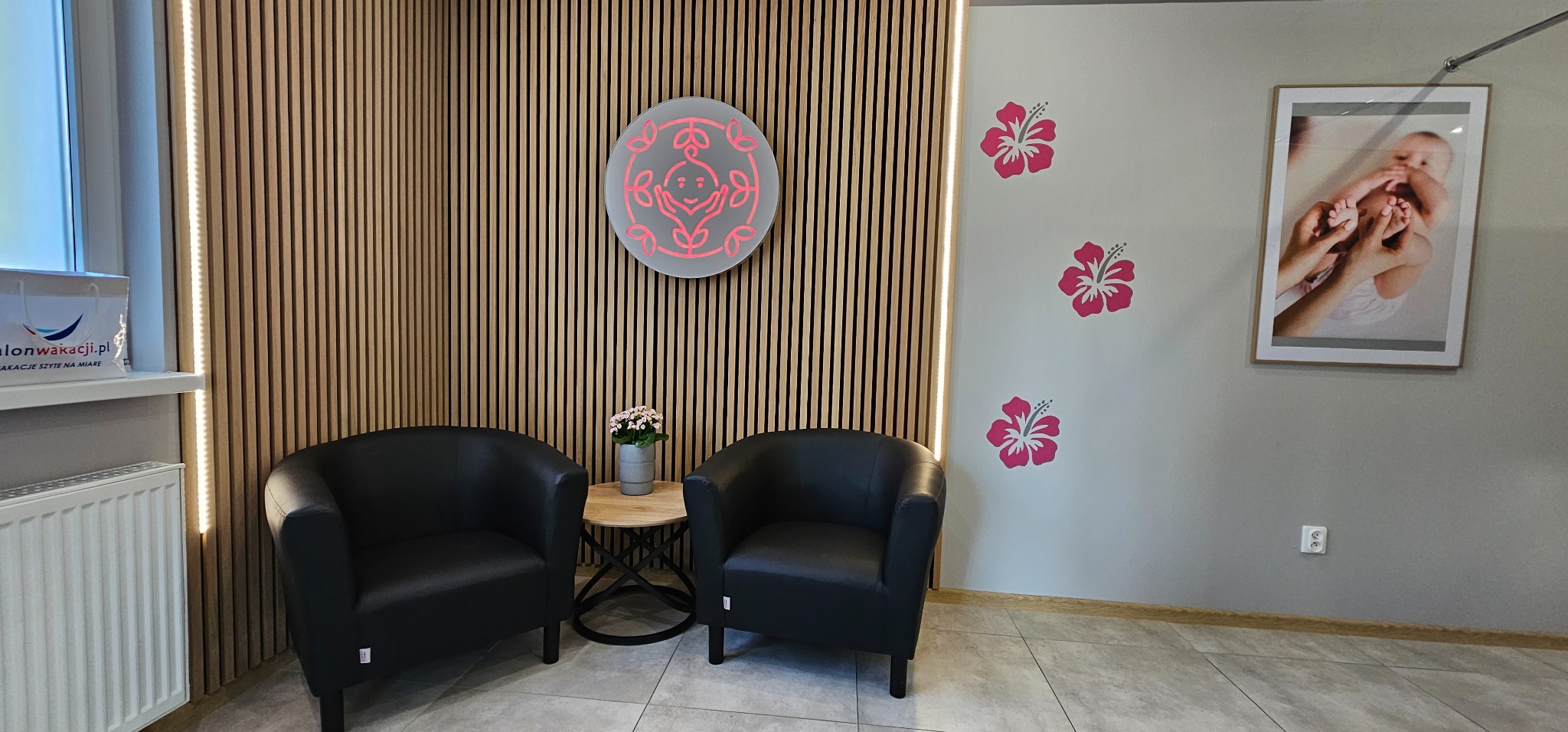 Poczekalnia: Dwa czarne fotele klubowe, okrągły stolik z kwiatami, drewniana ściana z podświetleniem LED, logo i grafika kwiatowa na ścianie, zdjęcie dziecka w ramce.