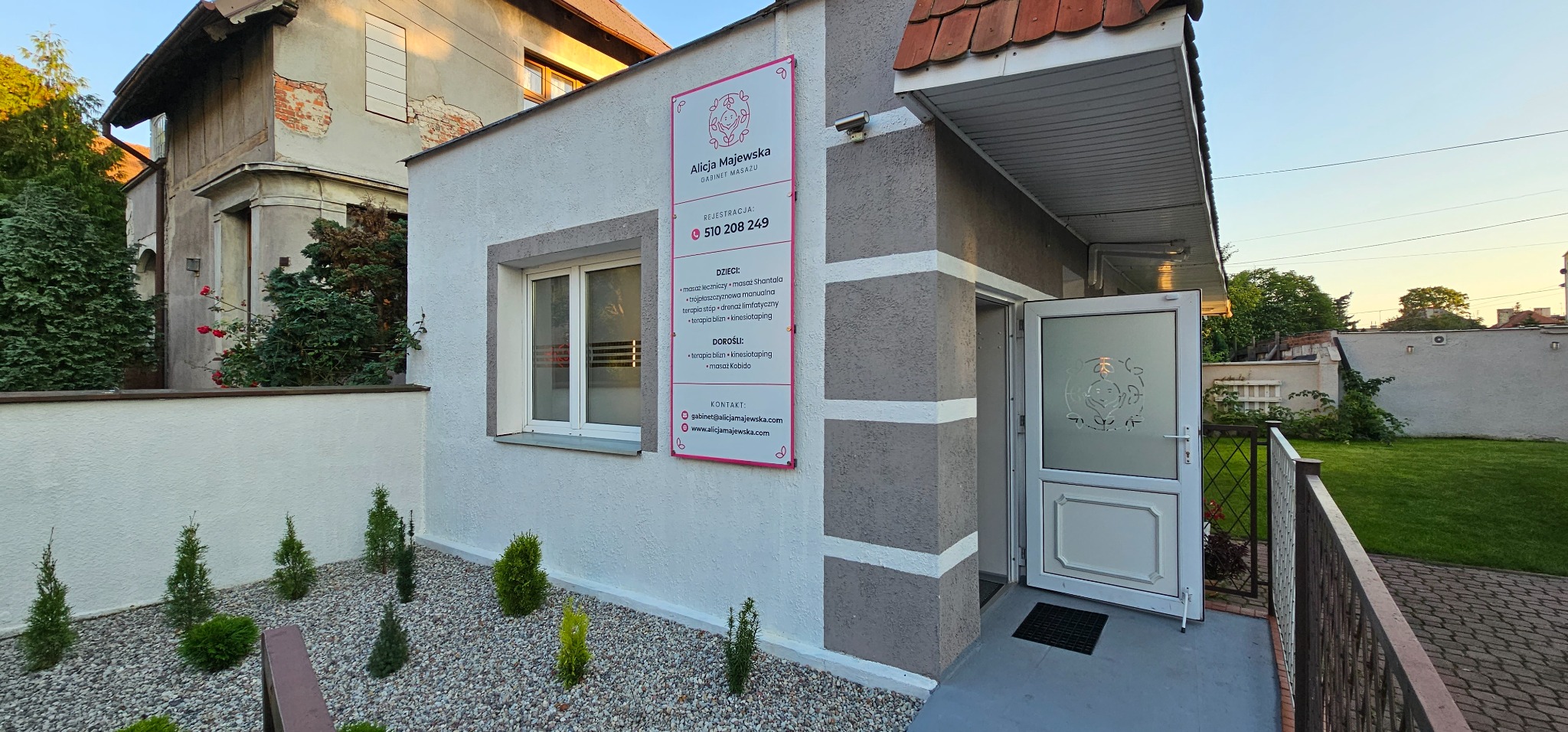 Gabinet masażu Alicja Majewska: wejście do budynku z tablicą informacyjną o ofercie dla dzieci i dorosłych, obejmującej masaże lecznicze, terapię blizn i kinesiotaping.