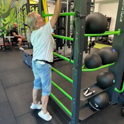 Ireneusz kwasniak - Kobieta w sportowym stroju przeciera wilgotną ściereczką metalową konstrukcję do ćwiczeń w siłowni; w tle widoczny mężczyzna siedzący na ławce.