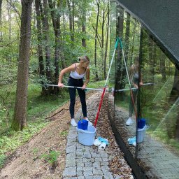 Ireneusz kwasniak - Kobieta w białym topie i czarnych spodniach czyści długą myjką okno nowoczesnego domu z dużymi przeszkleniami, otoczonego lasem; wiadro i ściereczki leżą na brukowanej ścieżce.