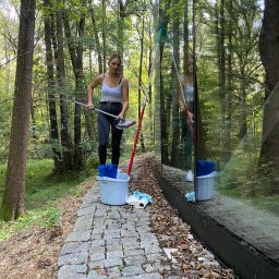 Ireneusz kwasniak - Młoda kobieta w trakcie mycia okien budynku otoczonego lasem, widoczny wiadro i akcesoria do mycia, odbicie w oknie.