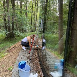 Ireneusz kwasniak - Kobieta w białym topie i czarnych spodniach myje okno w lesie za pomocą ściągaczki, widoczne narzędzia i wiadro Vileda, odbicie w oknie.