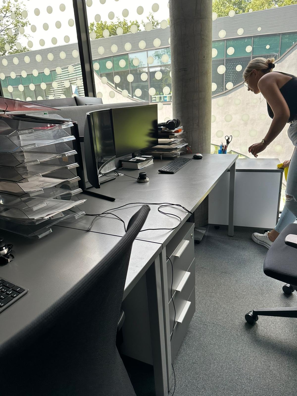 Kobieta w jeansach i białych butach czyści biurko w nowoczesnym biurze, widoczne liczne dokumenty w organizerach, monitor i okno z okrągłymi wzorami.