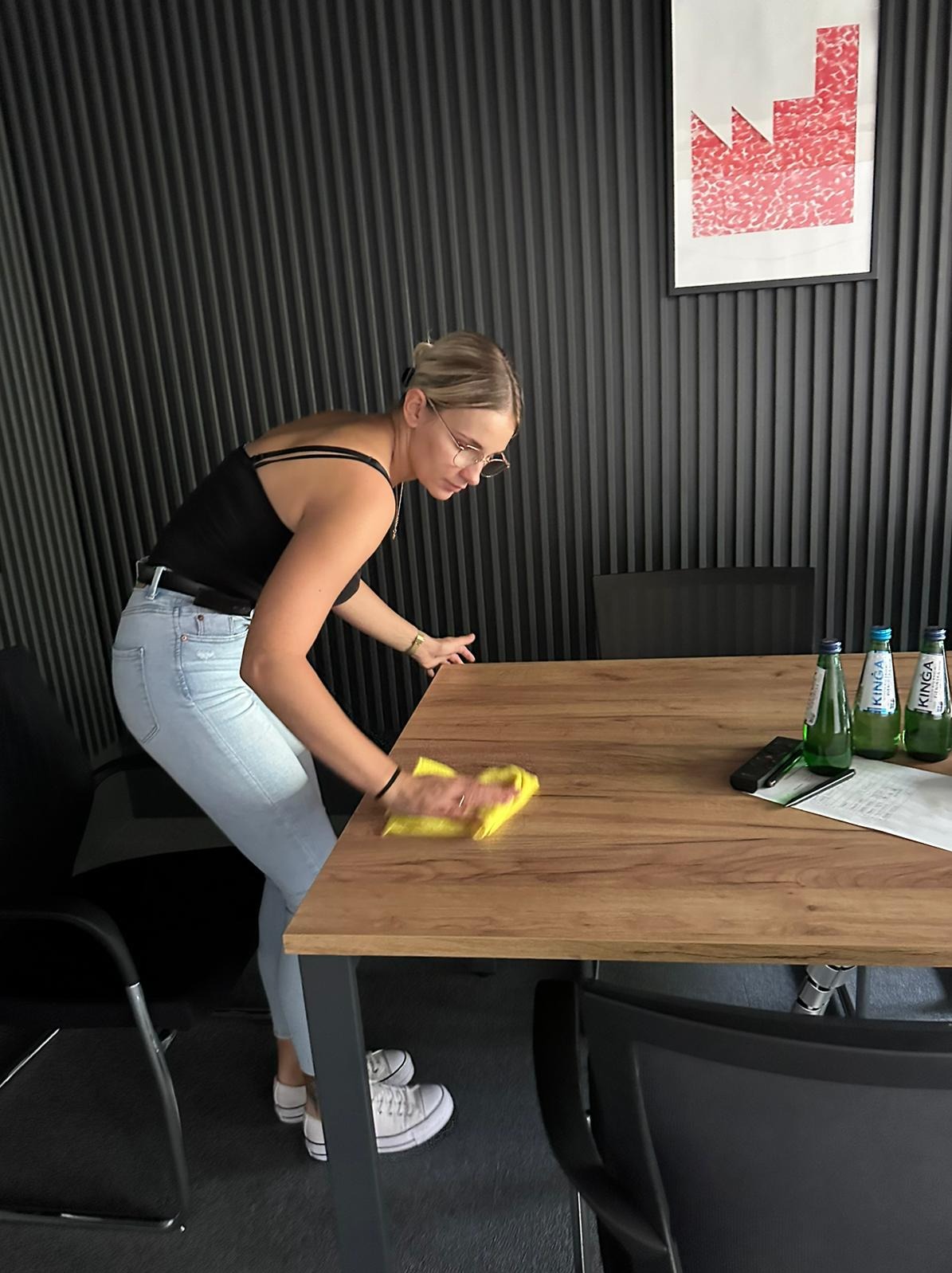 Kobieta w okularach przeciera żółtą ścierką drewniany stół w biurze, na którym stoją butelki wody Kinga, pilot i dokumenty, widoczne czarne krzesła i ściana w pionowe pasy z obrazem.