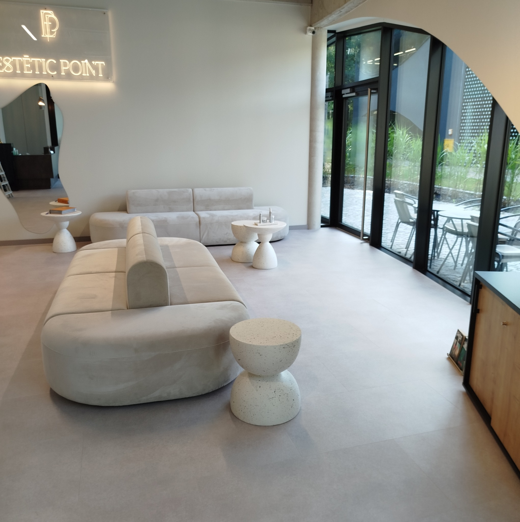 Jasne, minimalistyczne wnętrze poczekalni z sofami w odcieniach beżu i małymi stolikami o fakturze betonu, widok na zewnątrz przez duże okna.