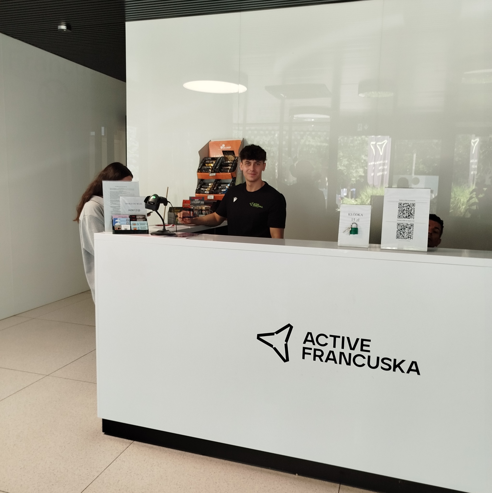 Recepcja w jasnym wnętrzu z pracownikiem za ladą, widoczne materiały informacyjne i produkty na sprzedaż