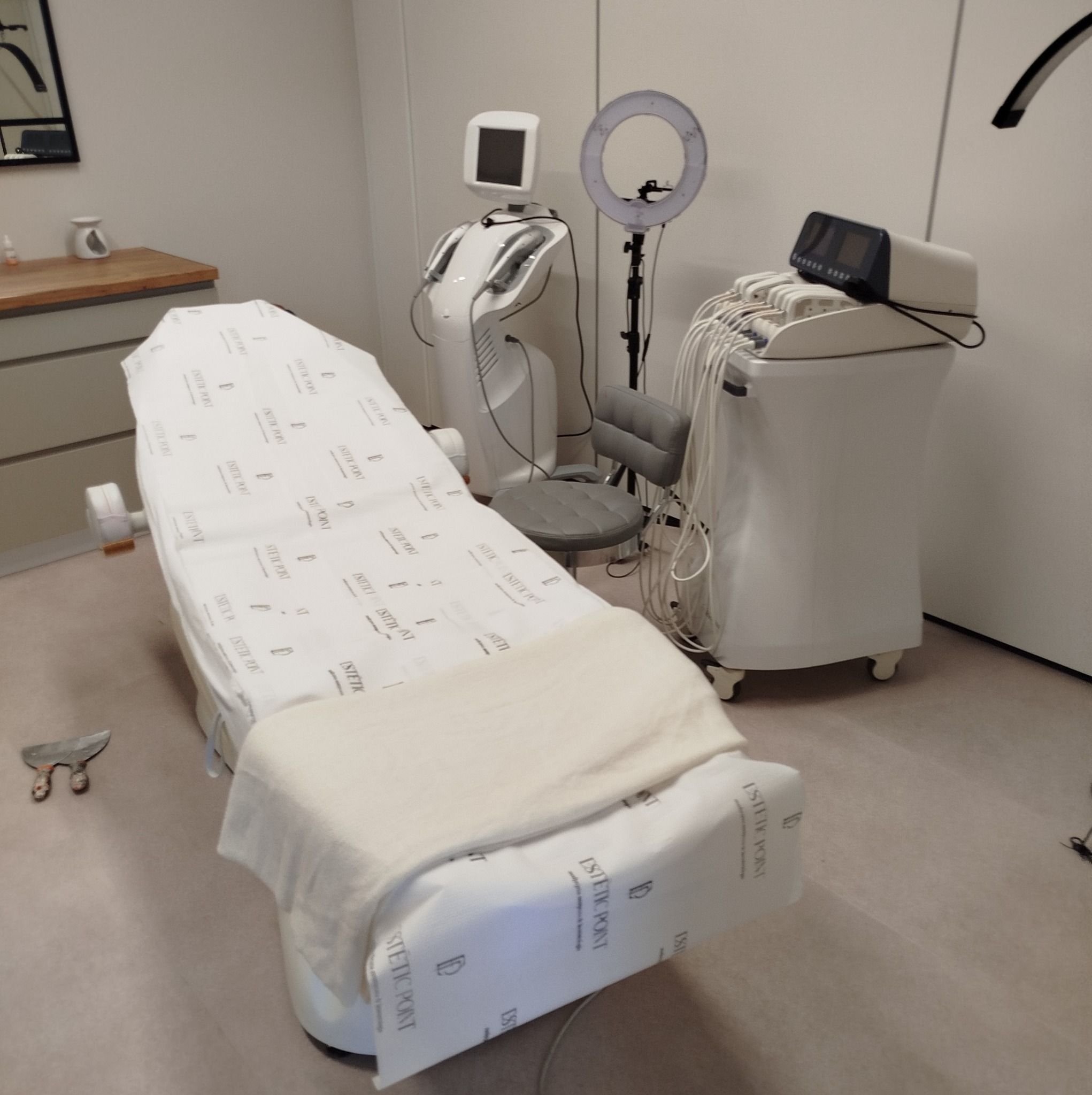Przygotowane stanowisko zabiegowe z leżanką przykrytą papierem z nadrukowanym logo, obok widoczne specjalistyczne urządzenia kosmetyczne, lampa pierścieniowa i krzesło