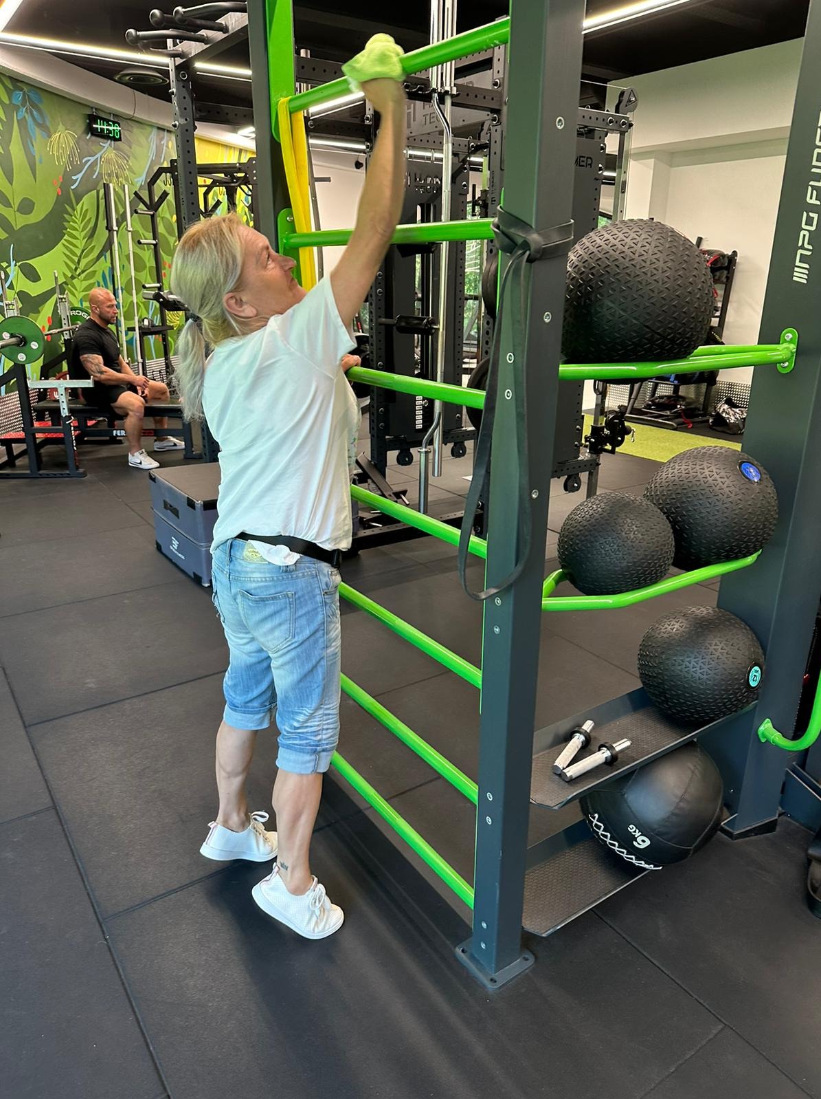 Kobieta w sportowym stroju przeciera wilgotną ściereczką metalową konstrukcję do ćwiczeń w siłowni; w tle widoczny mężczyzna siedzący na ławce.