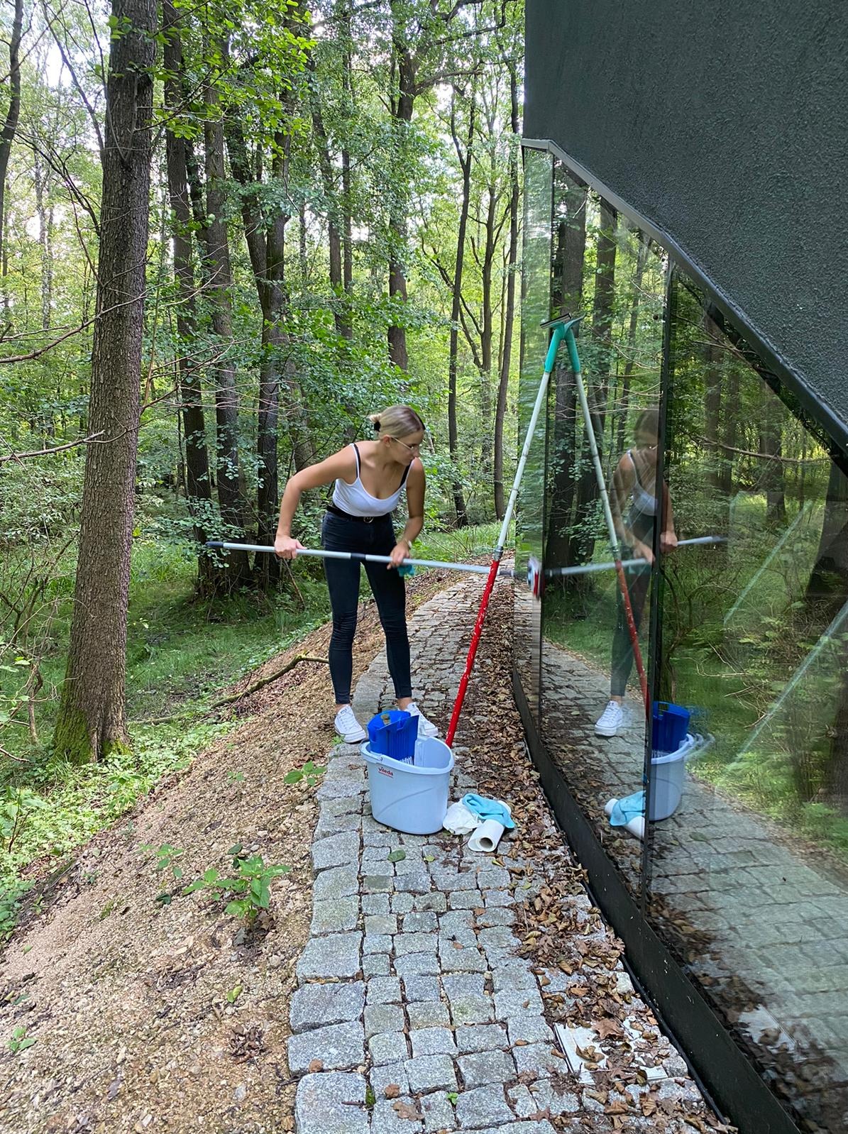 Kobieta w białym topie i czarnych spodniach czyści długą myjką okno nowoczesnego domu z dużymi przeszkleniami, otoczonego lasem; wiadro i ściereczki leżą na brukowanej ścieżce.
