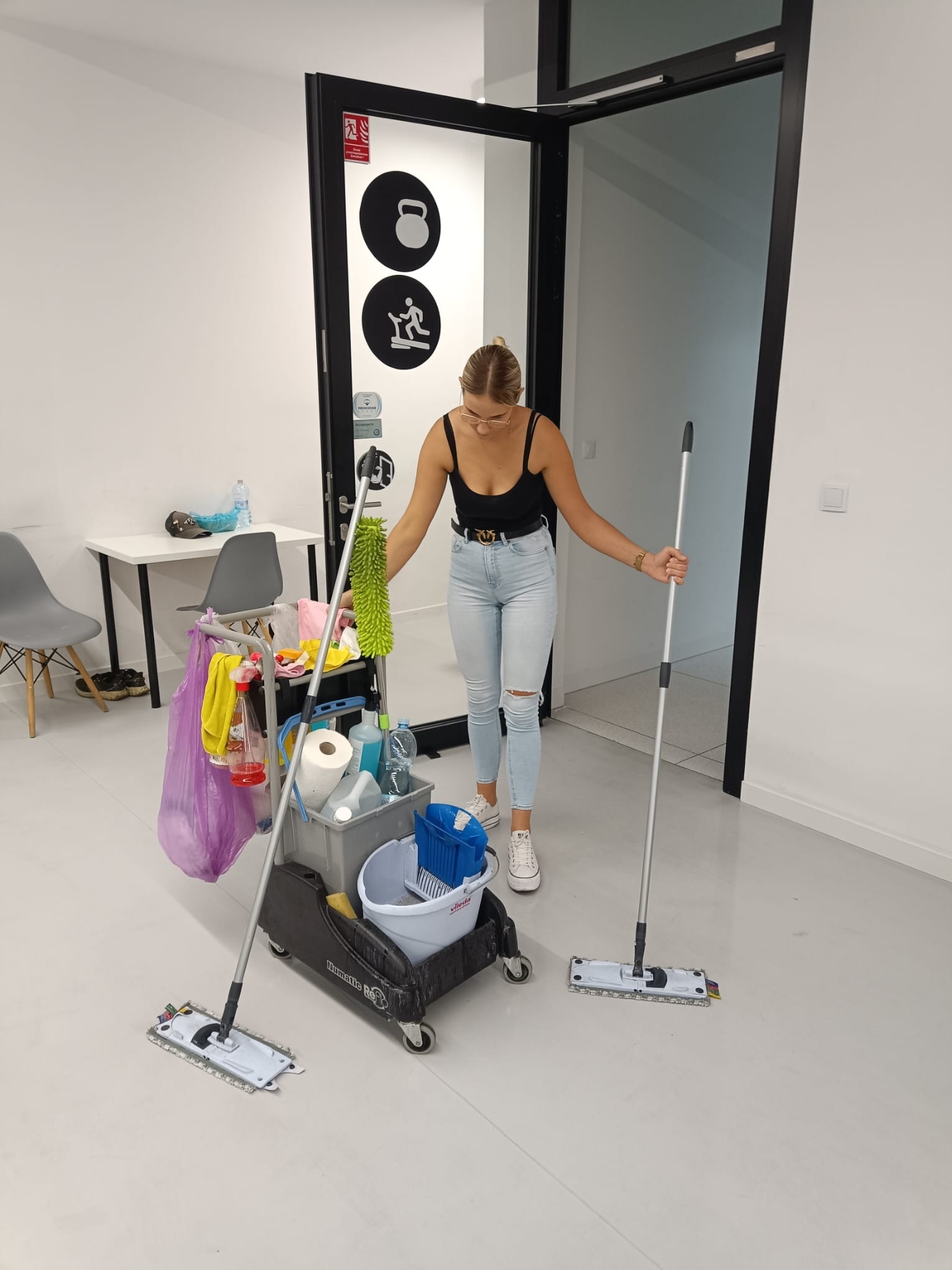Kobieta w jeansach i czarnym topie, z wózkiem pełnym środków czystości i mopami, w jasnym biurze z białymi ścianami i szarą podłogą, przygotowuje się do sprzątania.