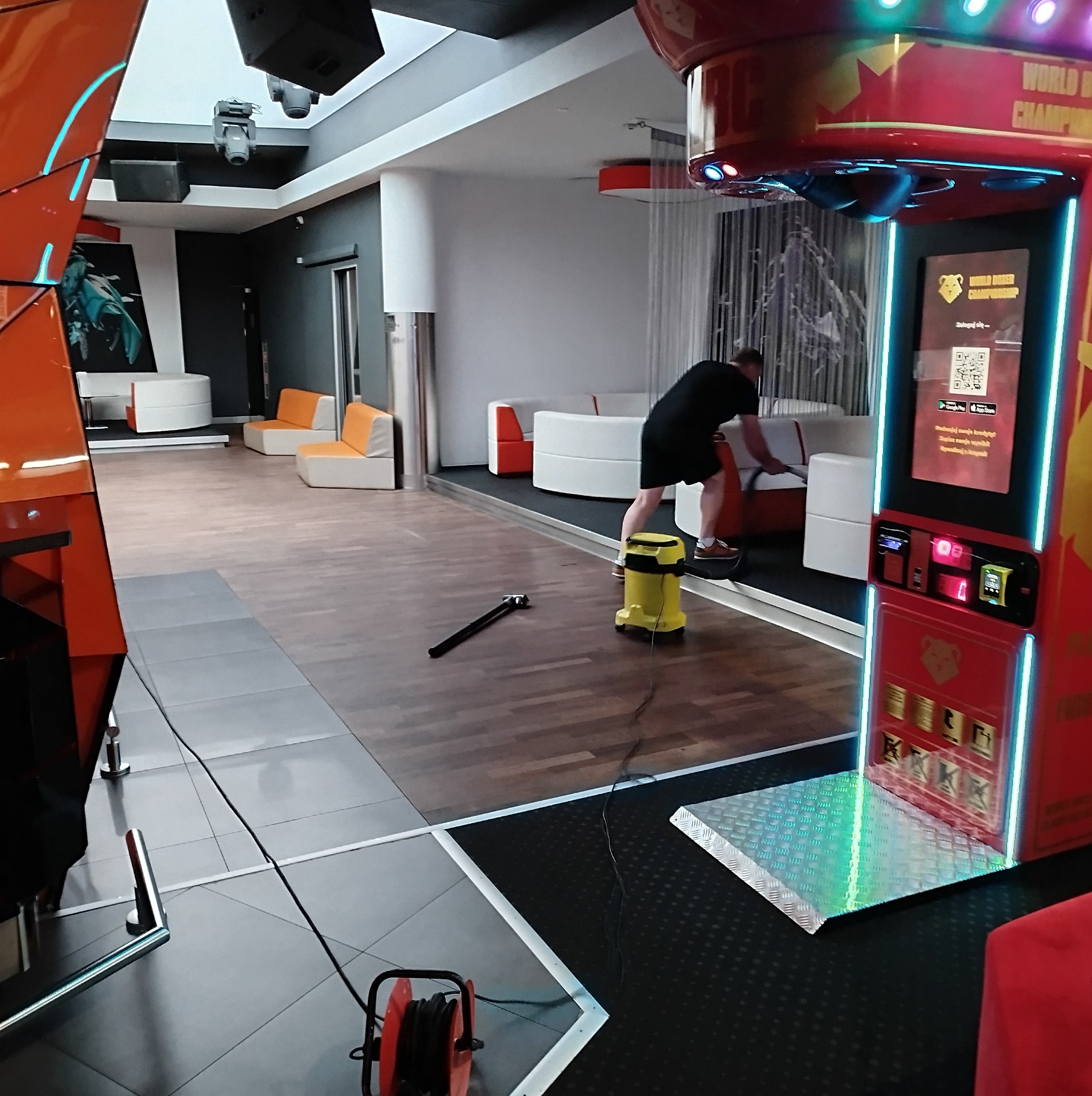 Pracownik w Katowicach odkurza podłogę w nowoczesnym pomieszczeniu rekreacyjnym, widoczny żółty odkurzacz przemysłowy i automat do gier.