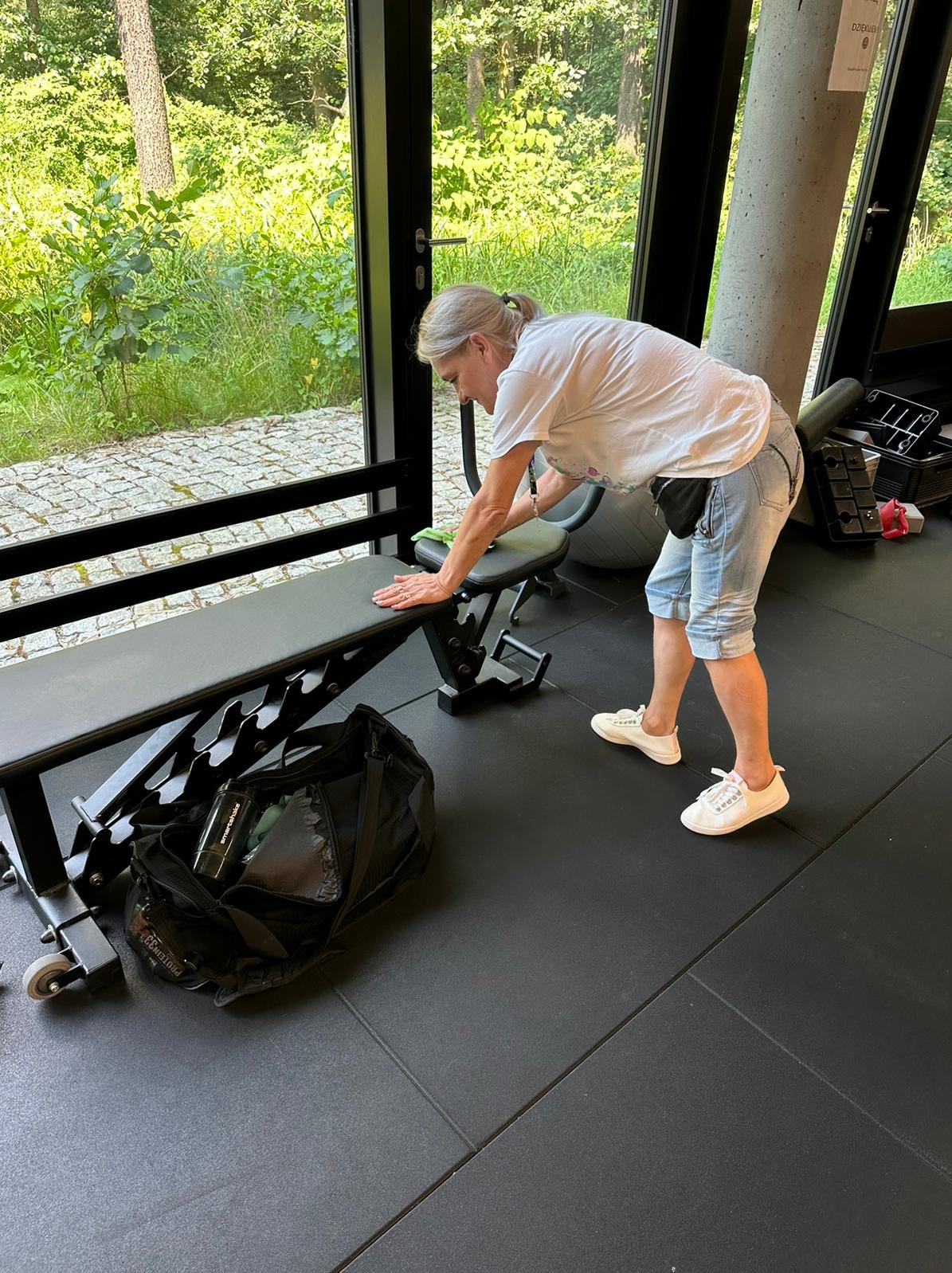 Kobieta w białej koszulce i jeansowych szortach przeciera zieloną ściereczką czarną ławkę do ćwiczeń w siłowni z widokiem na zieleń za oknem.