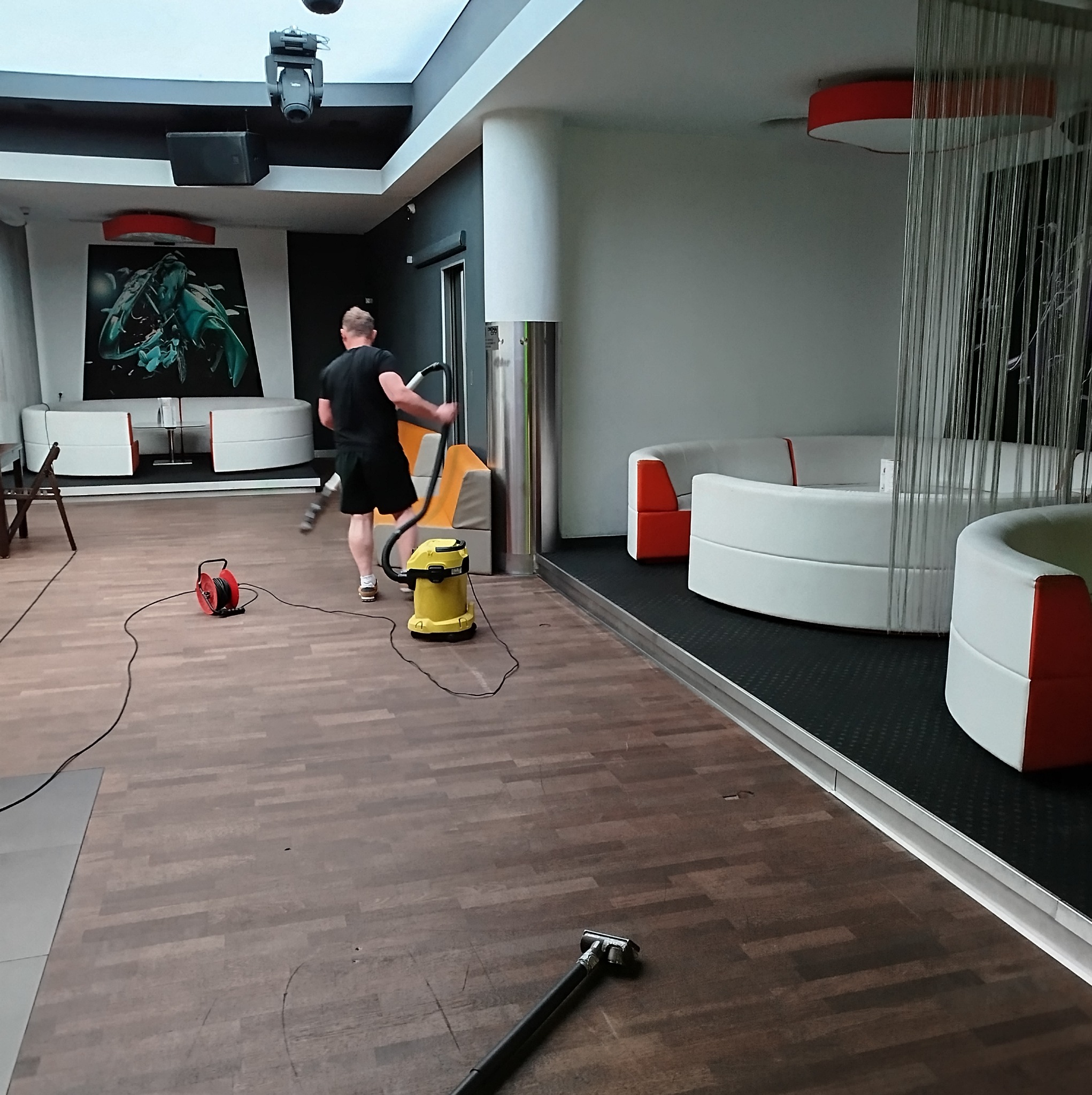 Mężczyzna odkurza podłogę w nowoczesnym klubie nocnym za pomocą odkurzacza przemysłowego; widoczne biało-pomarańczowe sofy, oświetlenie sceniczne i abstrakcyjny obraz na ścianie.