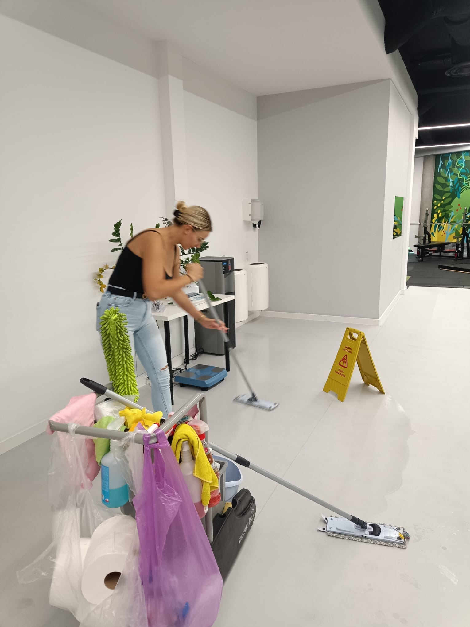 Pracownica w jeansach i czarnym topie myje podłogę mopem w jasnym biurze, w tle wózek z akcesoriami do sprzątania i znak ostrzegawczy 'Uwaga! Śliska podłoga'.