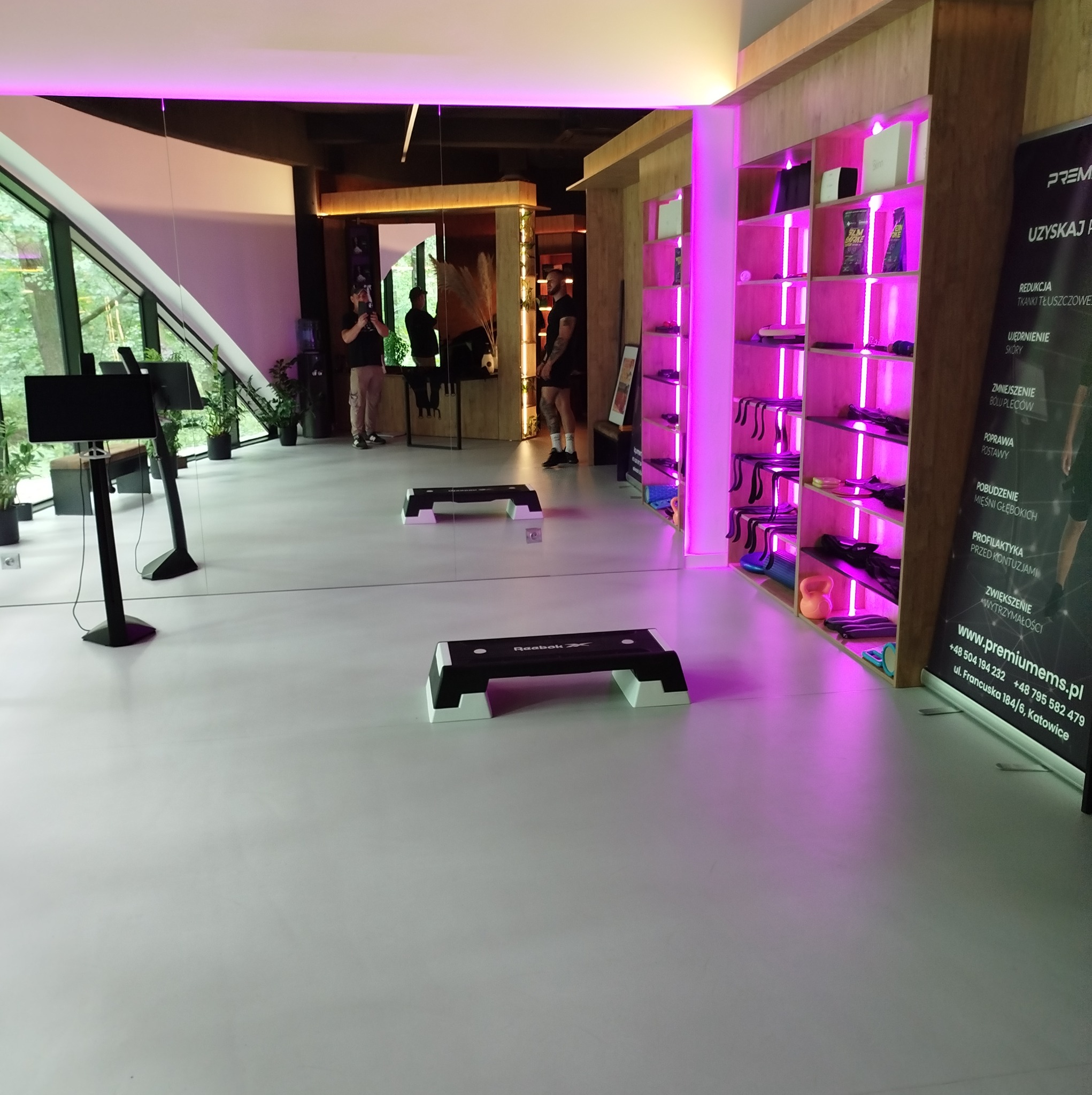 Jasne, przestronne wnętrze studia fitness z neonowym oświetleniem, sprzętem do ćwiczeń marki Reebok i lustrzaną ścianą odbijającą zielony krajobraz za oknem.