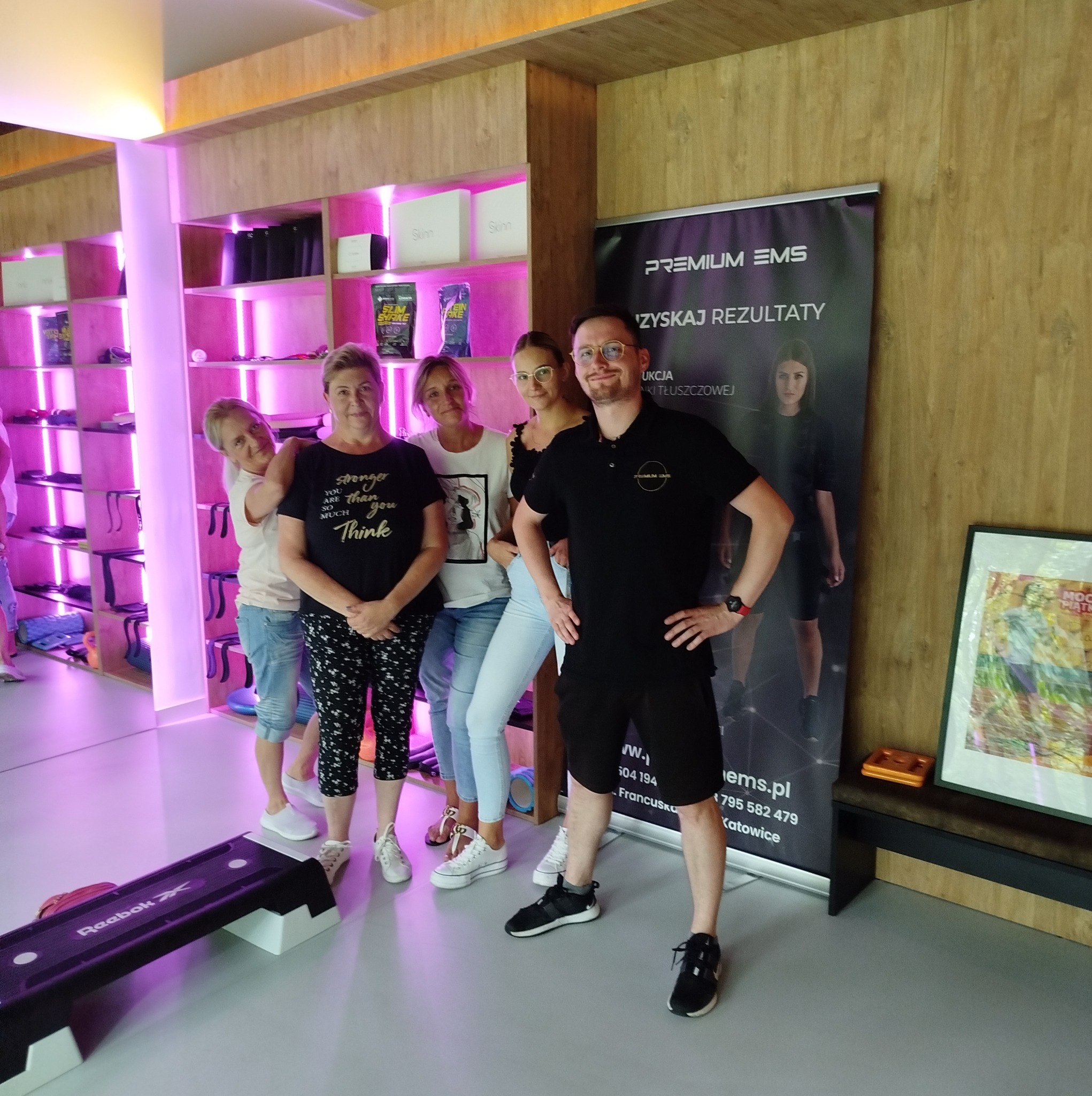 Grupa osób, w tym trener w czarnej koszulce z logo, pozuje w nowoczesnym studiu fitness z fioletowym oświetleniem, obok roll-upu reklamującego EMS i steppera Reebok.
