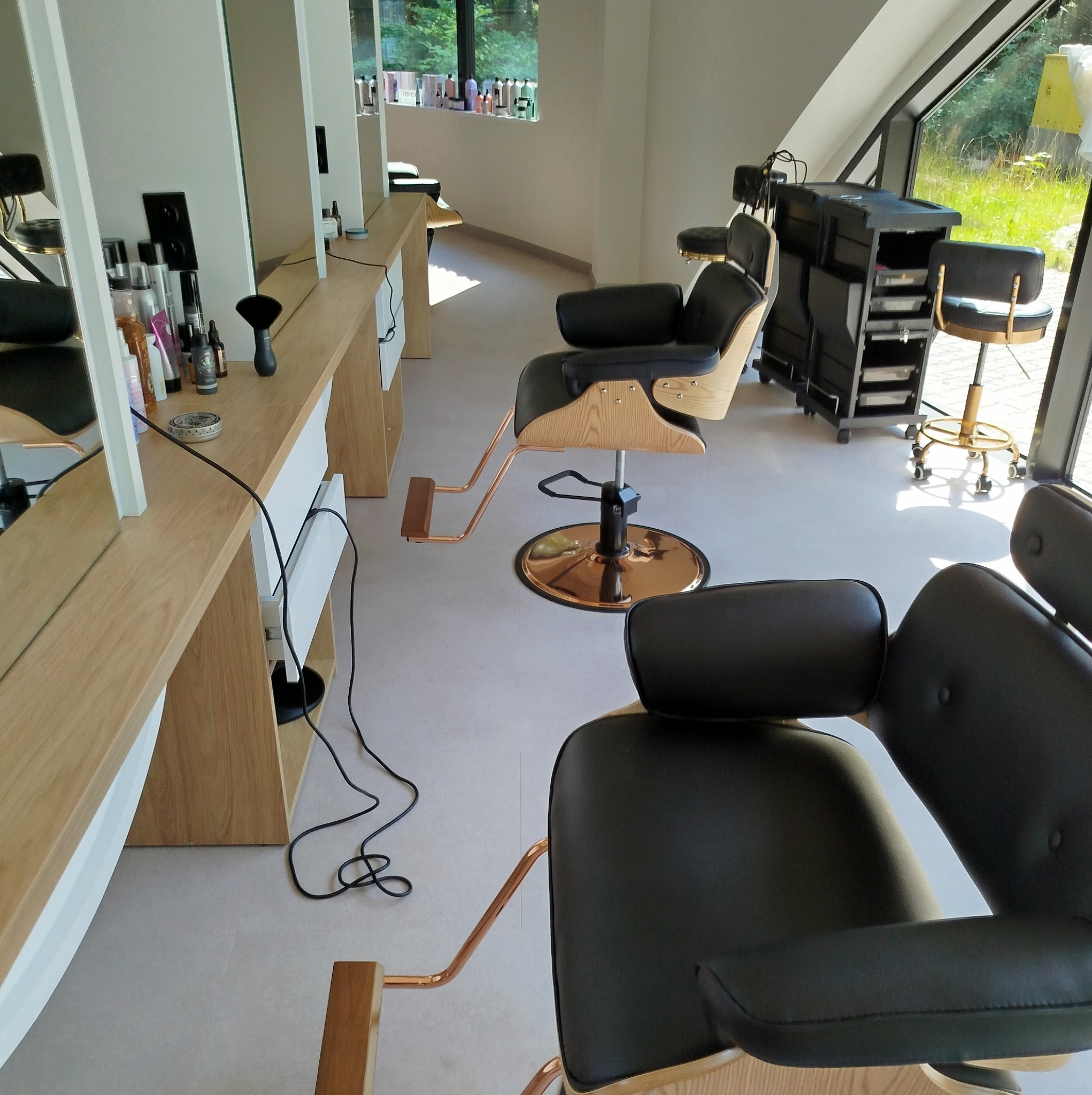 Jasne wnętrze salonu fryzjerskiego z drewnianymi stanowiskami i czarnymi fotelami fryzjerskimi z miedzianymi elementami, widok na okno z zielenią w tle.