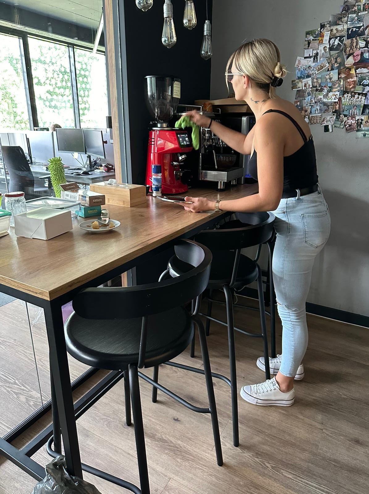 Kobieta w czarnym topie i jeansach czyści czerwoną maszynę do kawy w biurze, widoczne biurka w tle i ściana z kolażem zdjęć.