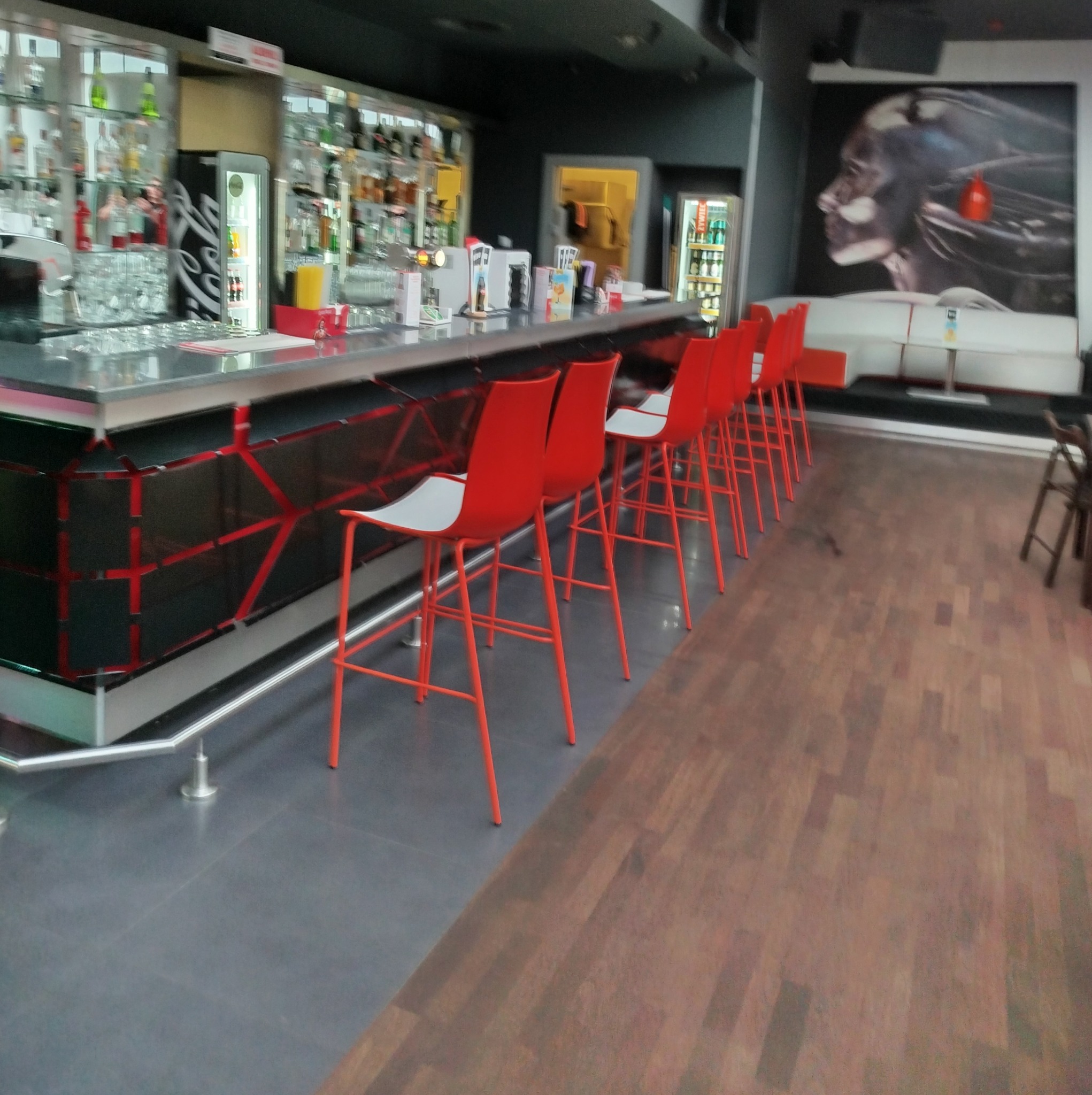 Wnętrze baru z rzędem czerwonych krzeseł barowych przy czarnym barze z podświetleniem, w tle regały z alkoholem i grafika na ścianie.