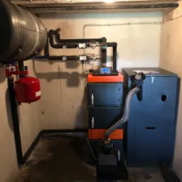 Kotłownia z kotłem na paliwo stałe, zbiornikiem akumulacyjnym i instalacją hydrauliczną w piwnicy, widoczne rury i zawory.