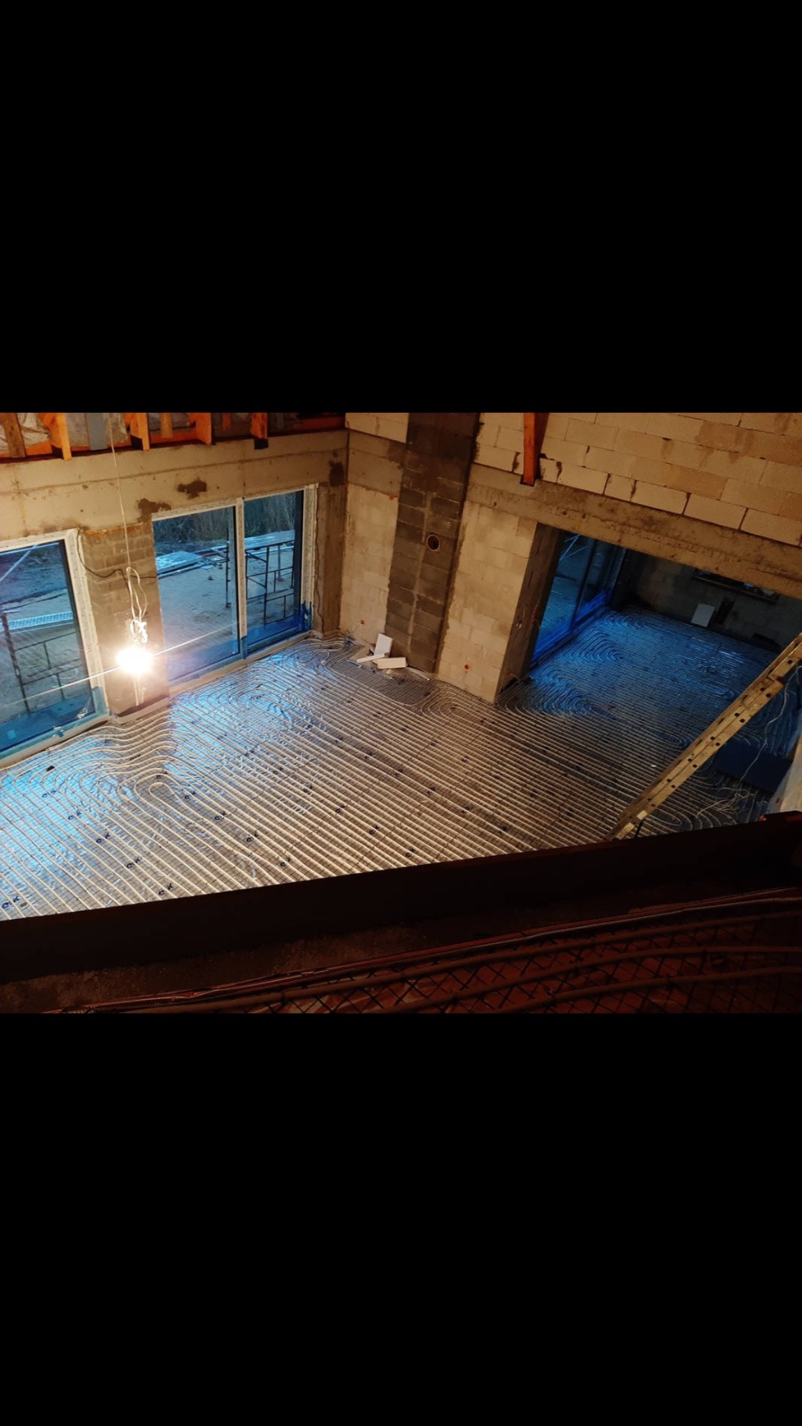 Widok z góry na rozłożoną instalację ogrzewania podłogowego w surowym stanie, rury ułożone spiralnie na folii izolacyjnej w pomieszczeniu z widocznymi ścianami z bloczków i oknami balkonowymi.