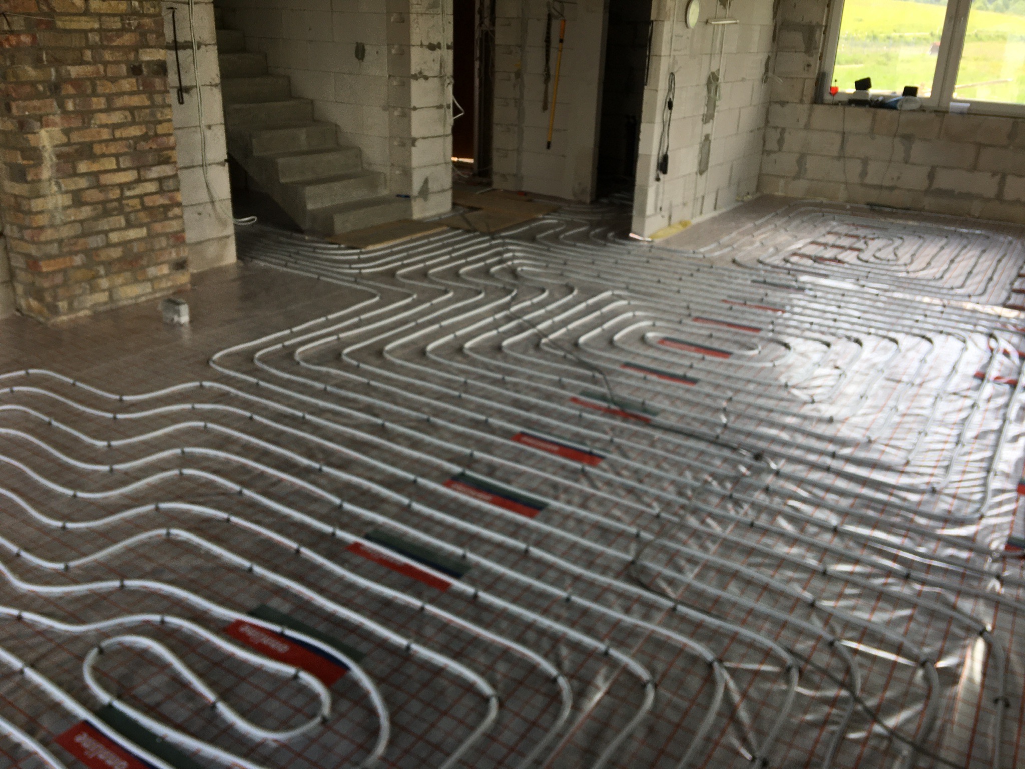 Rozłożone rury ogrzewania podłogowego w surowym wnętrzu domu w trakcie budowy, widoczne ściany z bloczków, schody betonowe i fragment ściany z cegły.