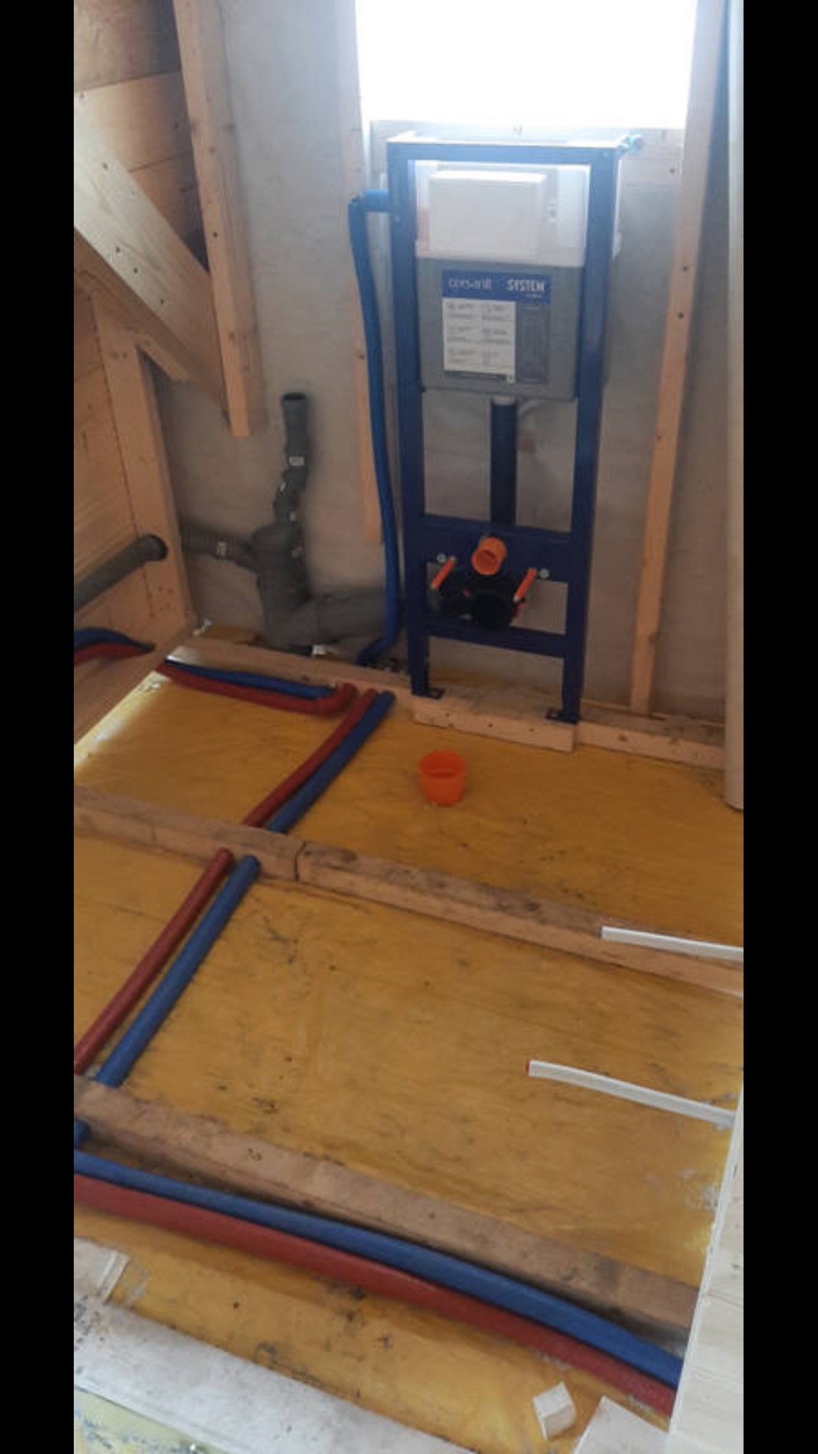 Montaż stelaża podtynkowego WC Geberit Duofix z niebieskimi rurami PEX w surowym wnętrzu z drewnianą konstrukcją szkieletową i żółtą folią izolacyjną na podłodze. Widoczna instalacja kanalizacyjna.