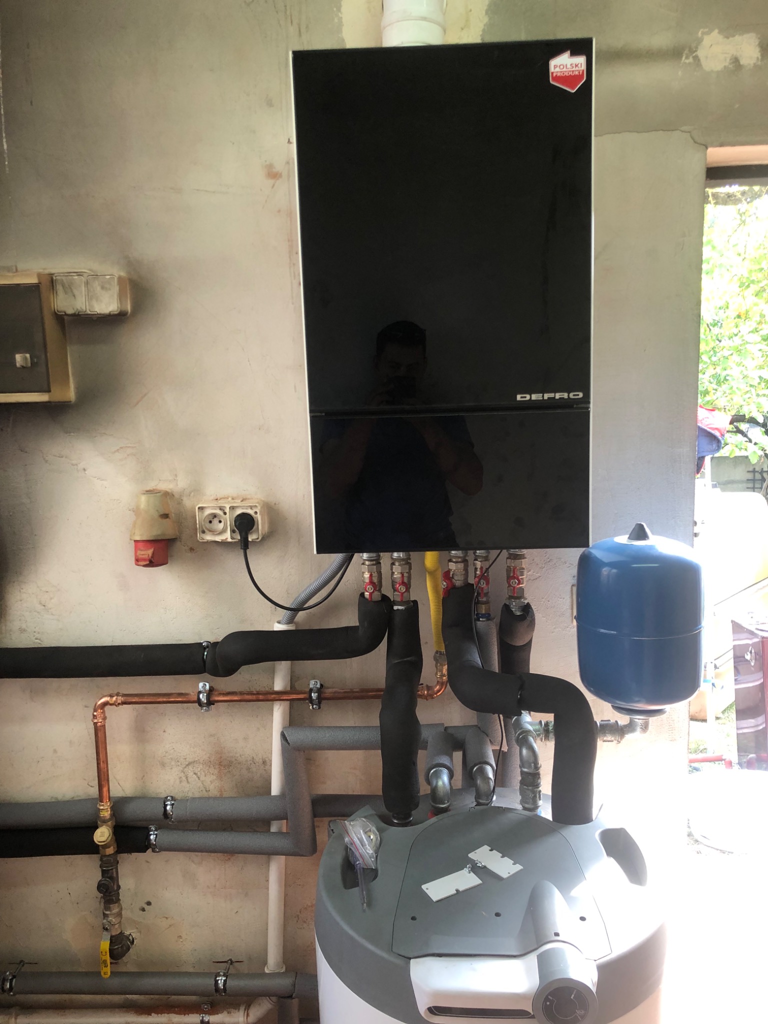 Czarny piec CO marki Defro z podłączonymi rurami miedzianymi i izolowanymi, zbiornik wyrównawczy niebieski, instalacja w surowym pomieszczeniu z widocznym oknem.